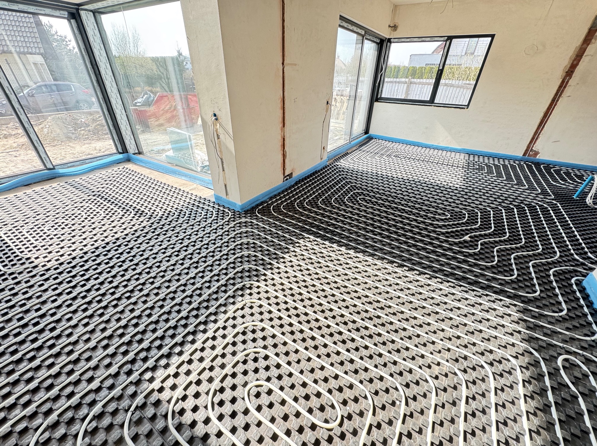 Białe rury ogrzewania podłogowego ułożone na czarnej macie w surowym wnętrzu z dużymi oknami. Instalacja przed zalaniem posadzki.