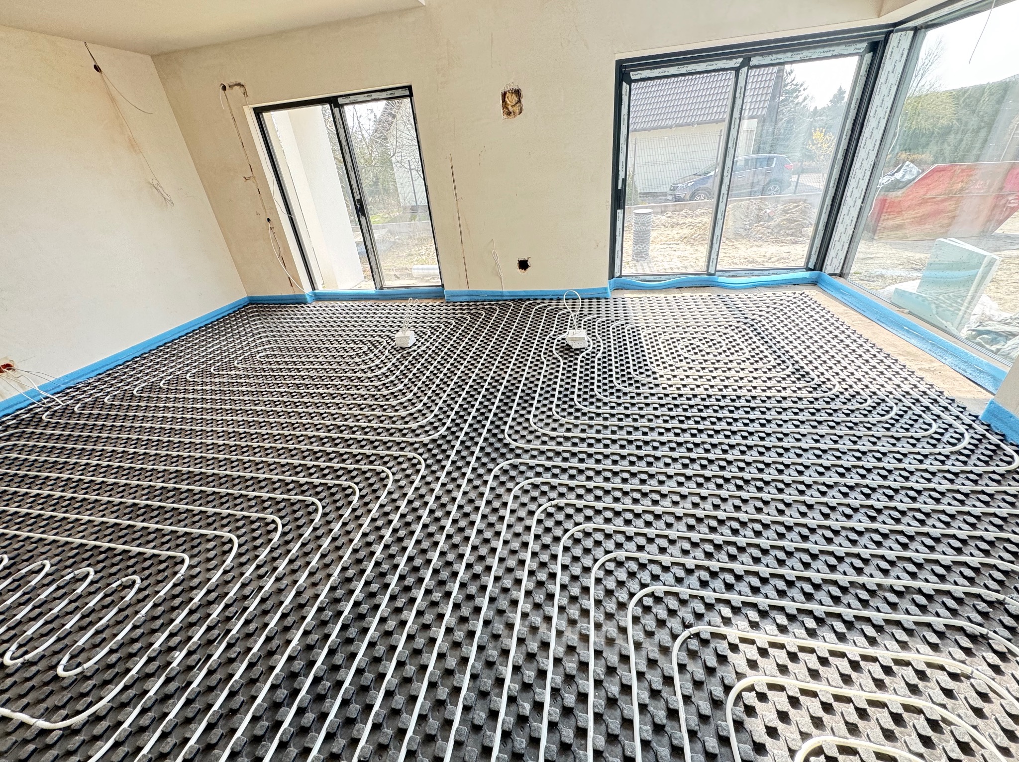Rozłożona instalacja ogrzewania podłogowego w surowym wnętrzu, widoczne białe rurki na czarnej macie, okna z widokiem na zewnątrz i fragmenty nieotynkowanych ścian.