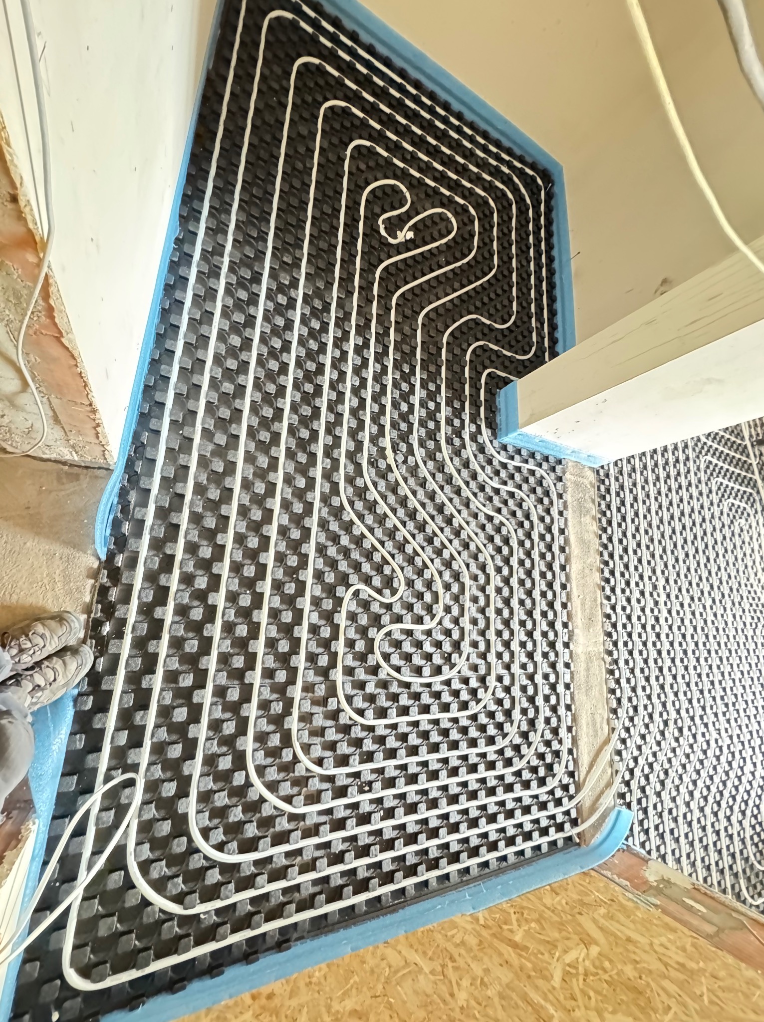 Instalacja ogrzewania podłogowego: białe rurki ułożone na czarnej macie z niebieską izolacją brzegową przy ścianie, widok z góry, częściowo widoczne buty instalatora.