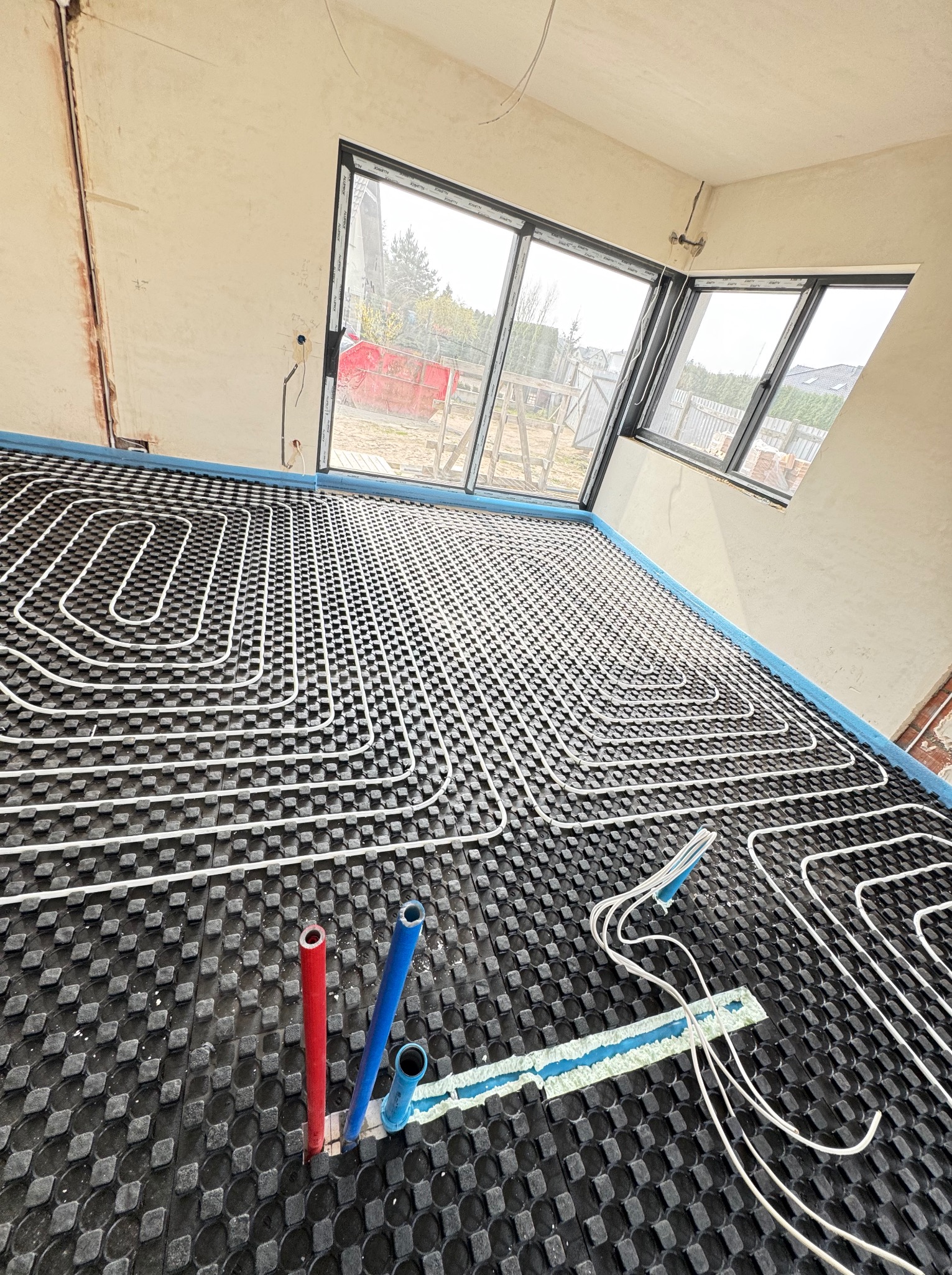 Instalacja ogrzewania podłogowego: białe rury ułożone na czarnej macie w nowym budynku, widoczne okno i wystające rury doprowadzające.