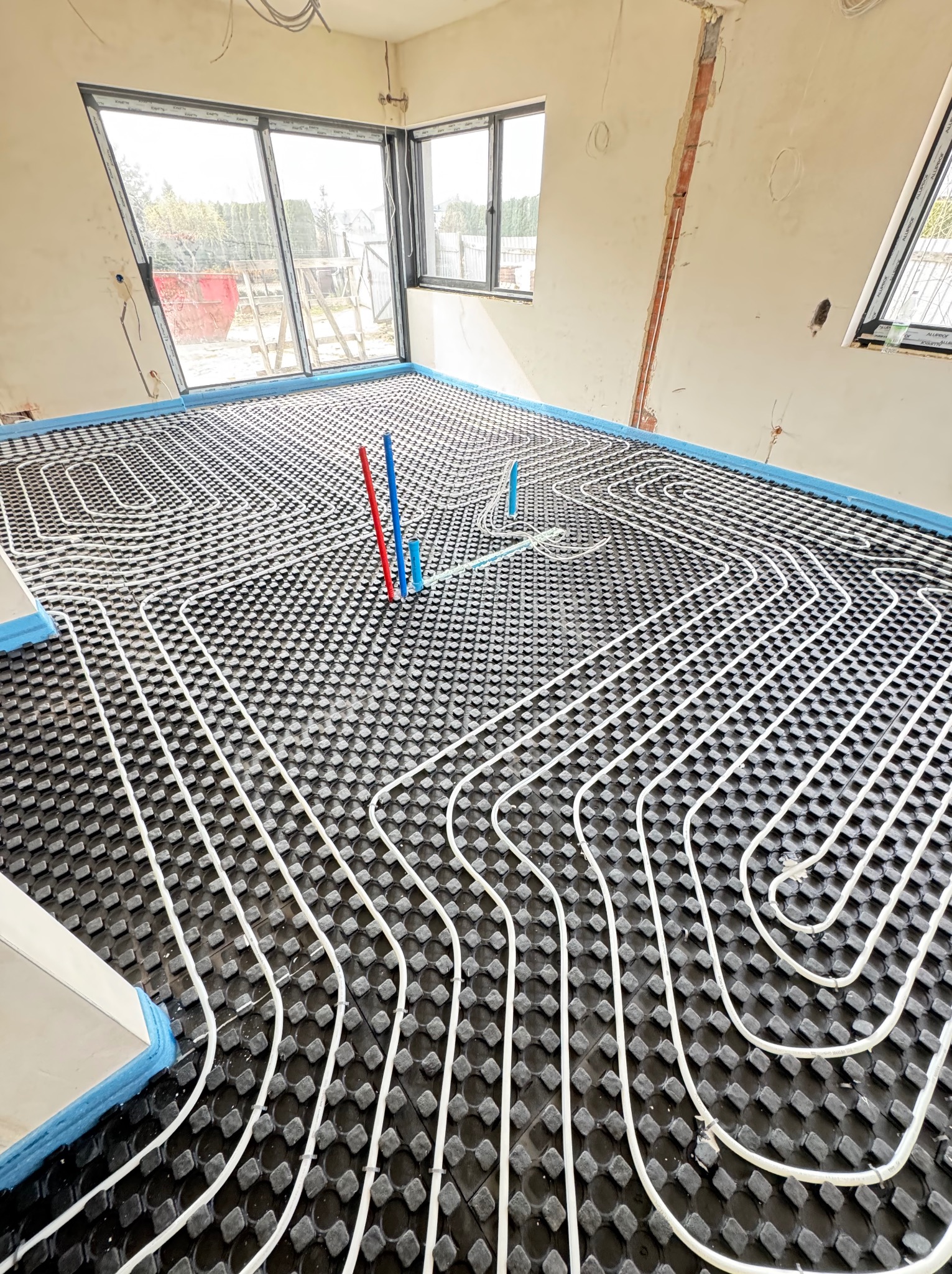Białe rury ogrzewania podłogowego ułożone na czarnej macie w surowym wnętrzu, z widocznymi pionami instalacji w kolorach niebieskim i czerwonym.