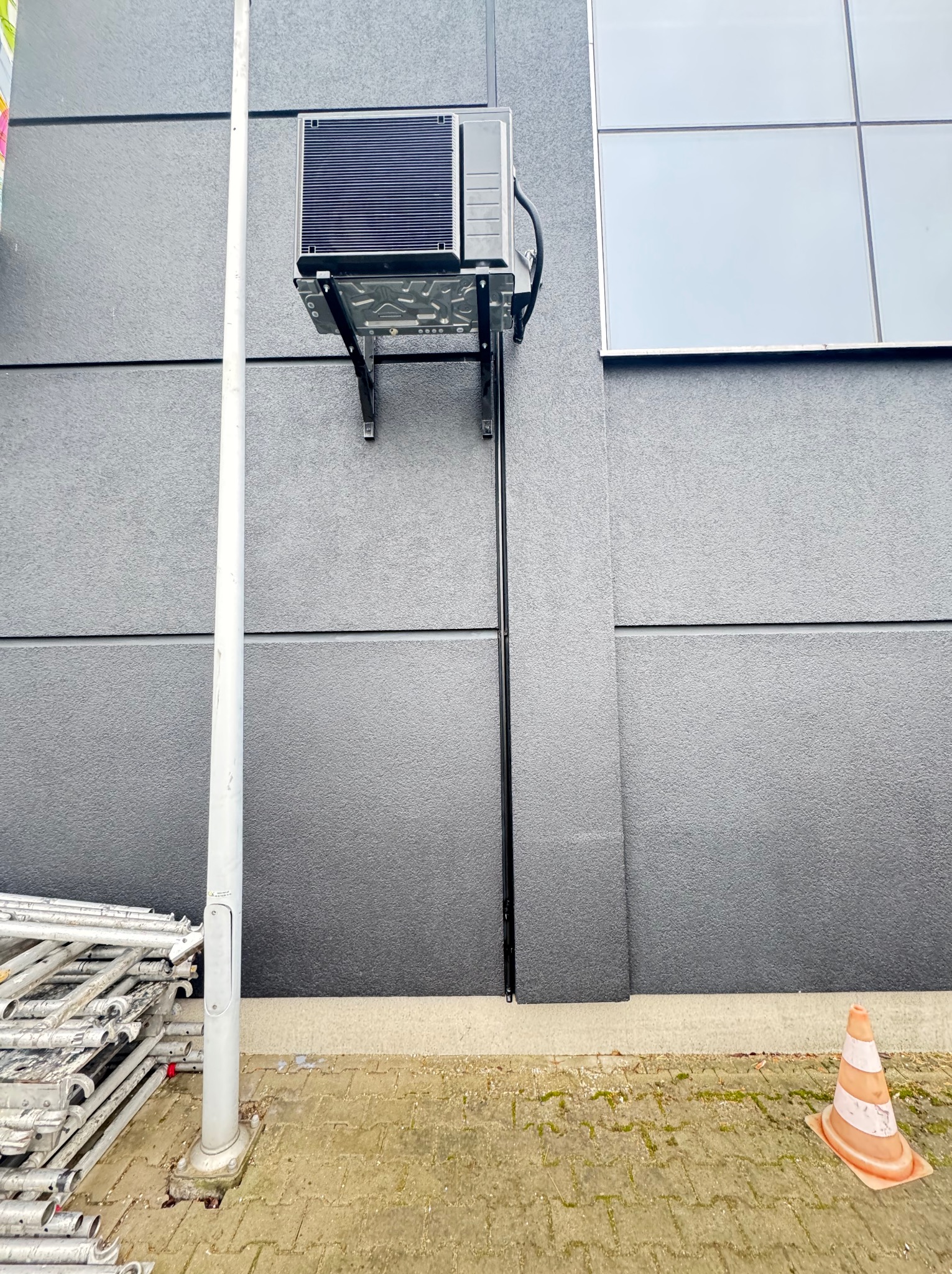 Zewnętrzny montaż klimatyzatora na szarej elewacji budynku, widoczne rusztowania i pachołek drogowy. Kąt ujęcia z dołu podkreśla wysokość instalacji.