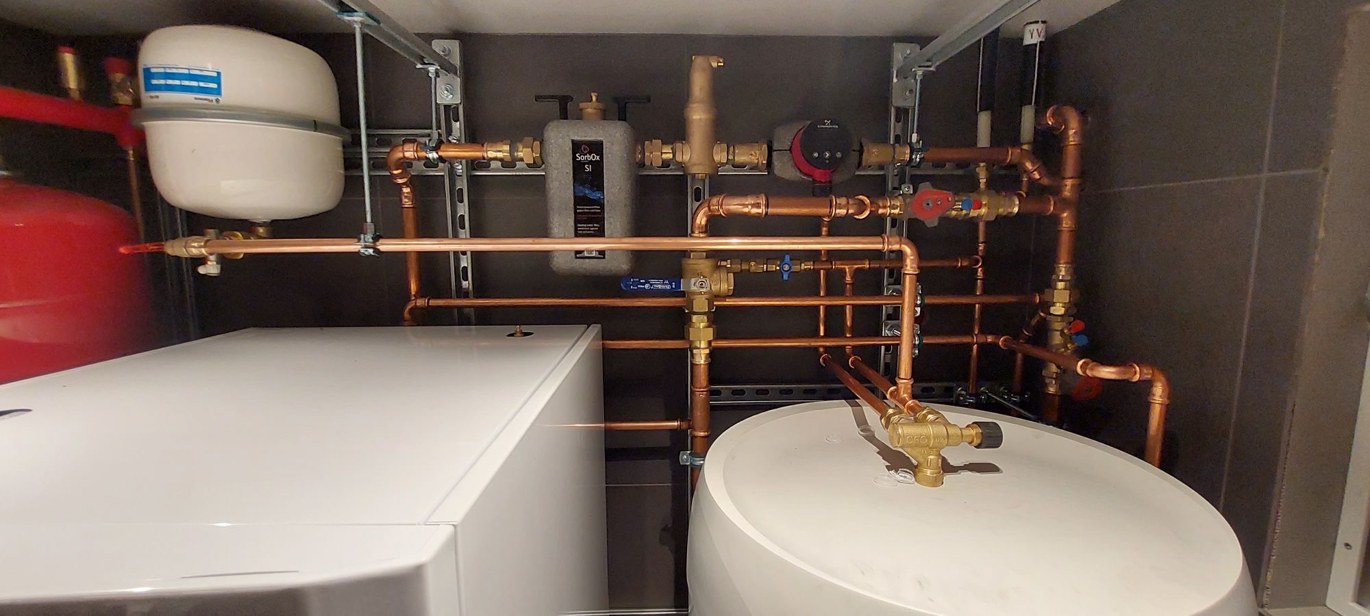 Kompleksowa instalacja grzewcza w kotłowni, widoczne miedziane rury, zawory, filtry, pompy obiegowe oraz zbiorniki wyrównawcze i ekspansyjne, zamontowane na szynach montażowych w pomieszczeniu...