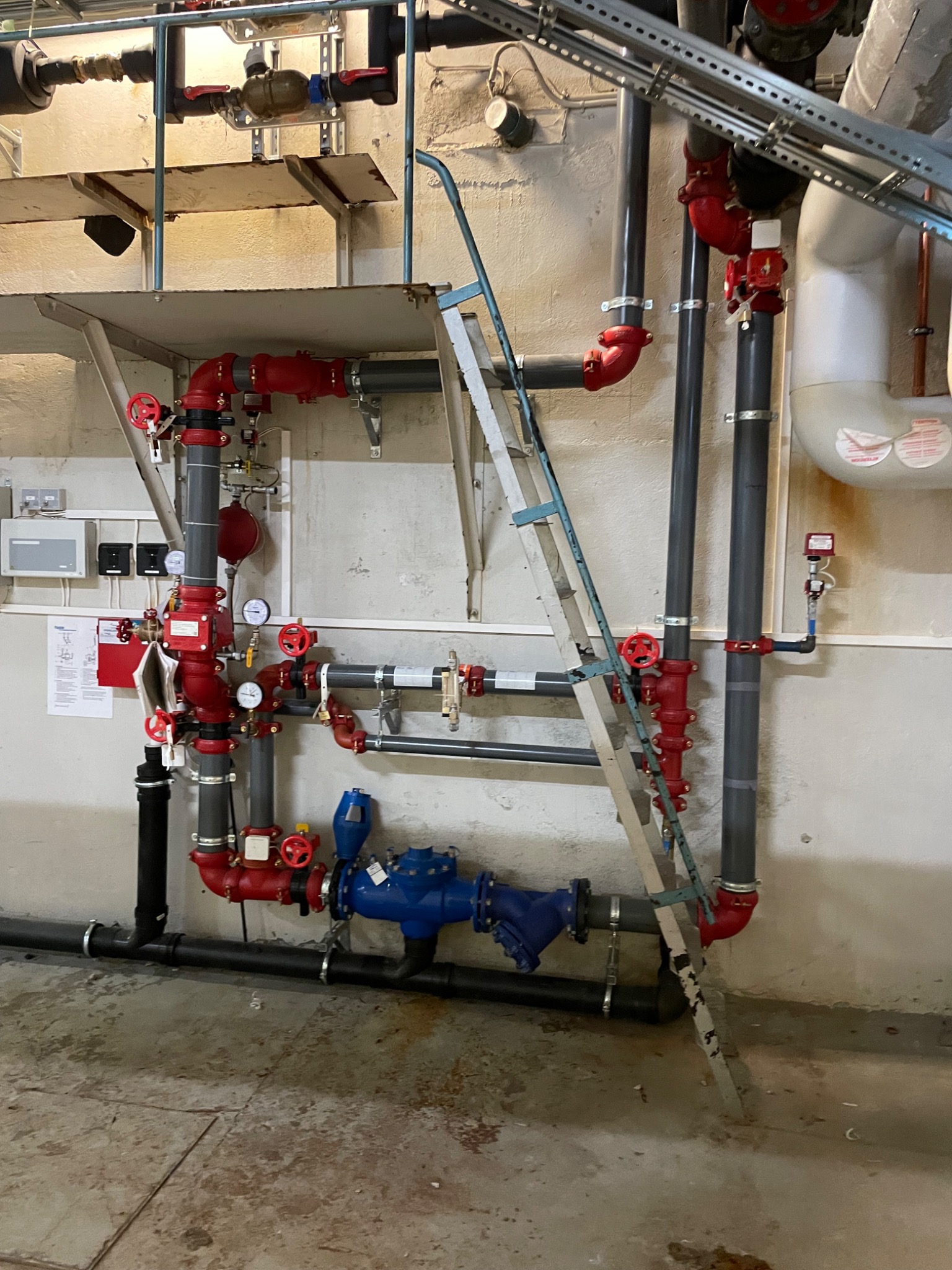 Złożona instalacja rur w kolorach czerwonym, czarnym i niebieskim, z licznymi zaworami i manometrami, częściowo zasłonięta przez aluminiową drabinę opartą o ścianę. Widoczne fragmenty sufitu...