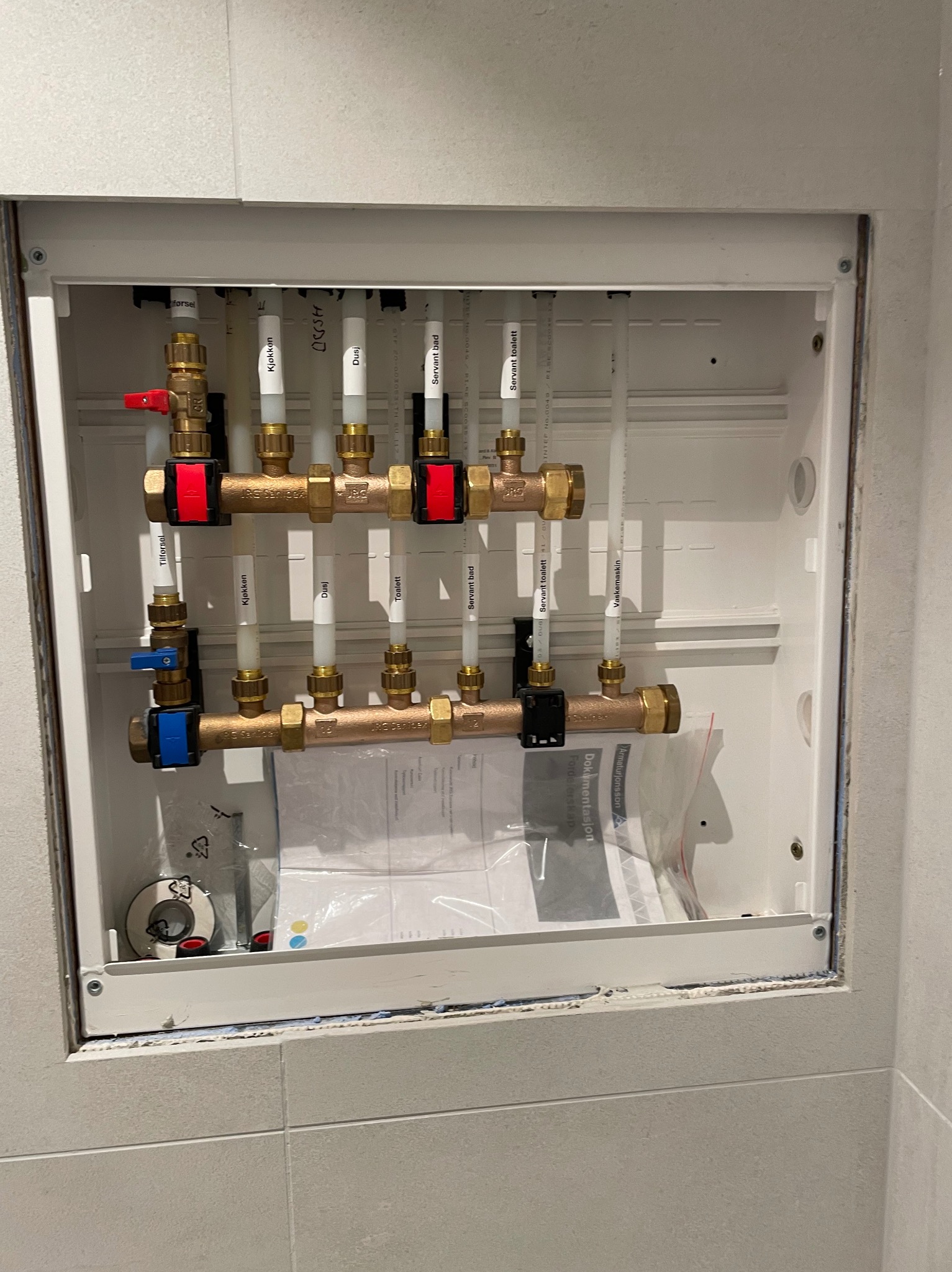Rozdzielacz instalacji wodnej w szafce wnękowej, z rurami oznaczonymi nazwami pomieszczeń (kuchnia, łazienka), zawory z czerwonymi i niebieskimi uchwytami, widoczne dokumenty instalacyjne wewnątrz...