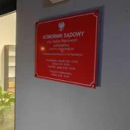 Czerwona tablica informacyjna na ścianie z napisem 'Komornik Sądowy przy Sądzie Rejonowym w Raciborzu' oraz godzinami urzędowania, widoczna obok drzwi z rośliną w tle.