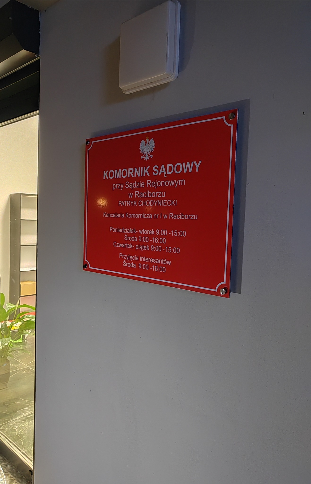 Czerwona tablica informacyjna na ścianie z napisem 'Komornik Sądowy przy Sądzie Rejonowym w Raciborzu' oraz godzinami urzędowania, widoczna obok drzwi z rośliną w tle.