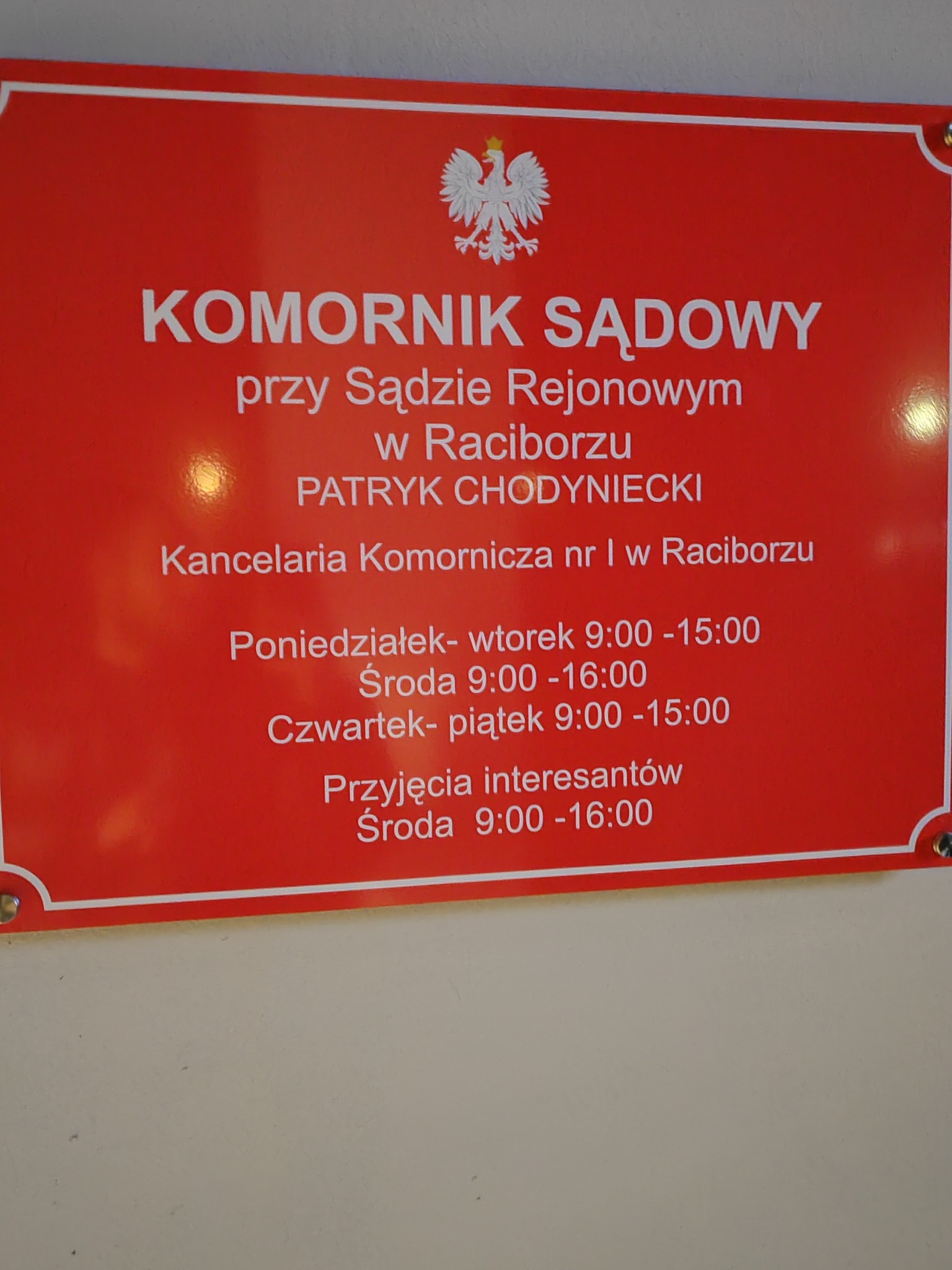 Czerwona tablica informacyjna z białym napisem: Komornik Sądowy przy Sądzie Rejonowym w Raciborzu, Patryk Chodyniecki, Kancelaria Komornicza nr I w Raciborzu, oraz godziny przyjęć.