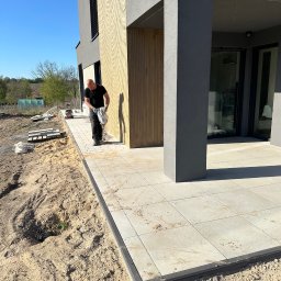 Dawid Wójciński - Układanie jasnej kostki brukowej na tarasie nowoczesnego domu. Brukarz w trakcie pracy, widoczna zagęszczarka i przygotowane płyty. Słoneczny dzień.