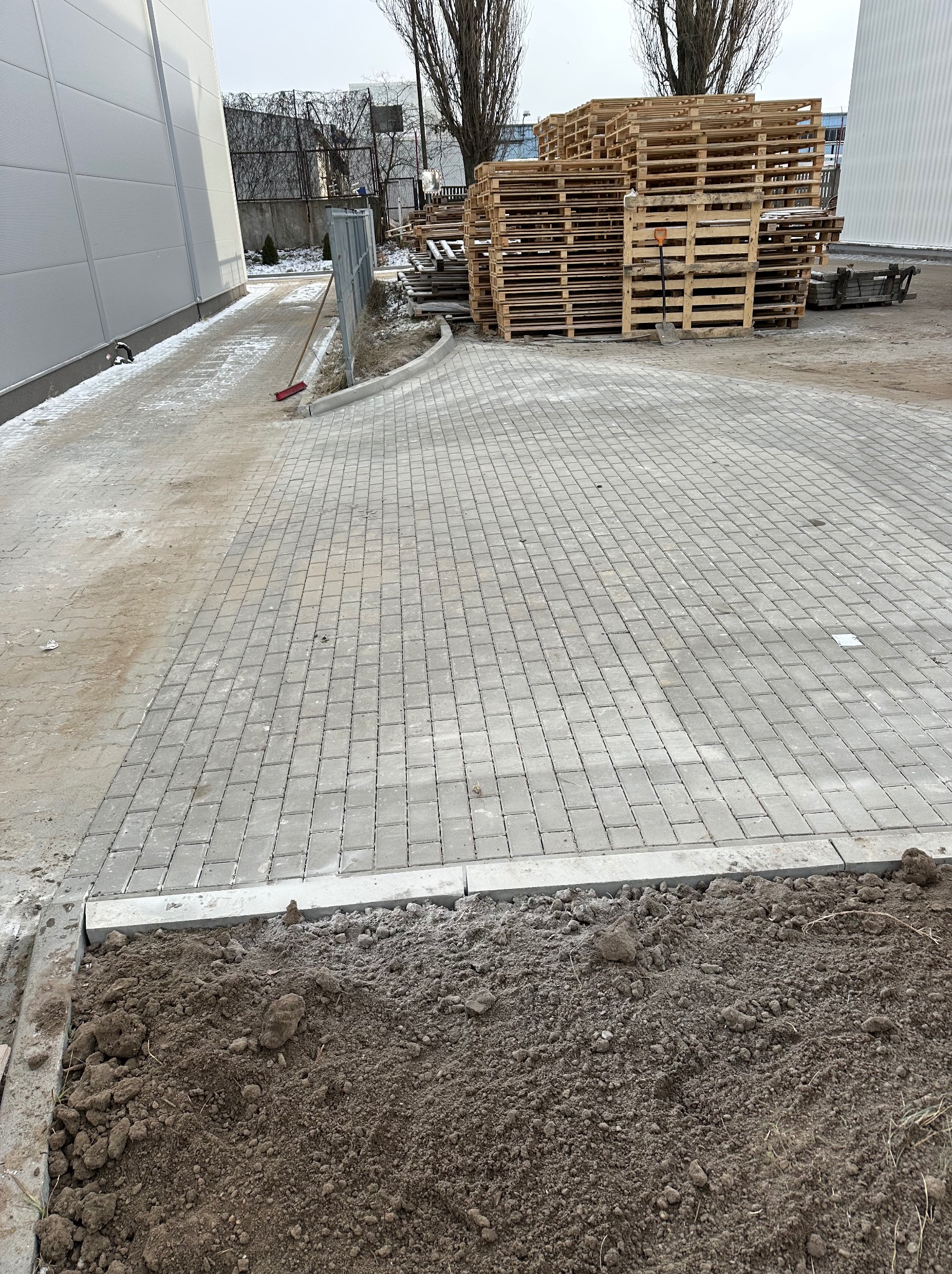 Świeżo ułożona szara kostka brukowa na posesji, z widocznymi stertami drewnianych palet w tle i fragmentem niezabudowanego terenu z ziemią w pierwszym planie.