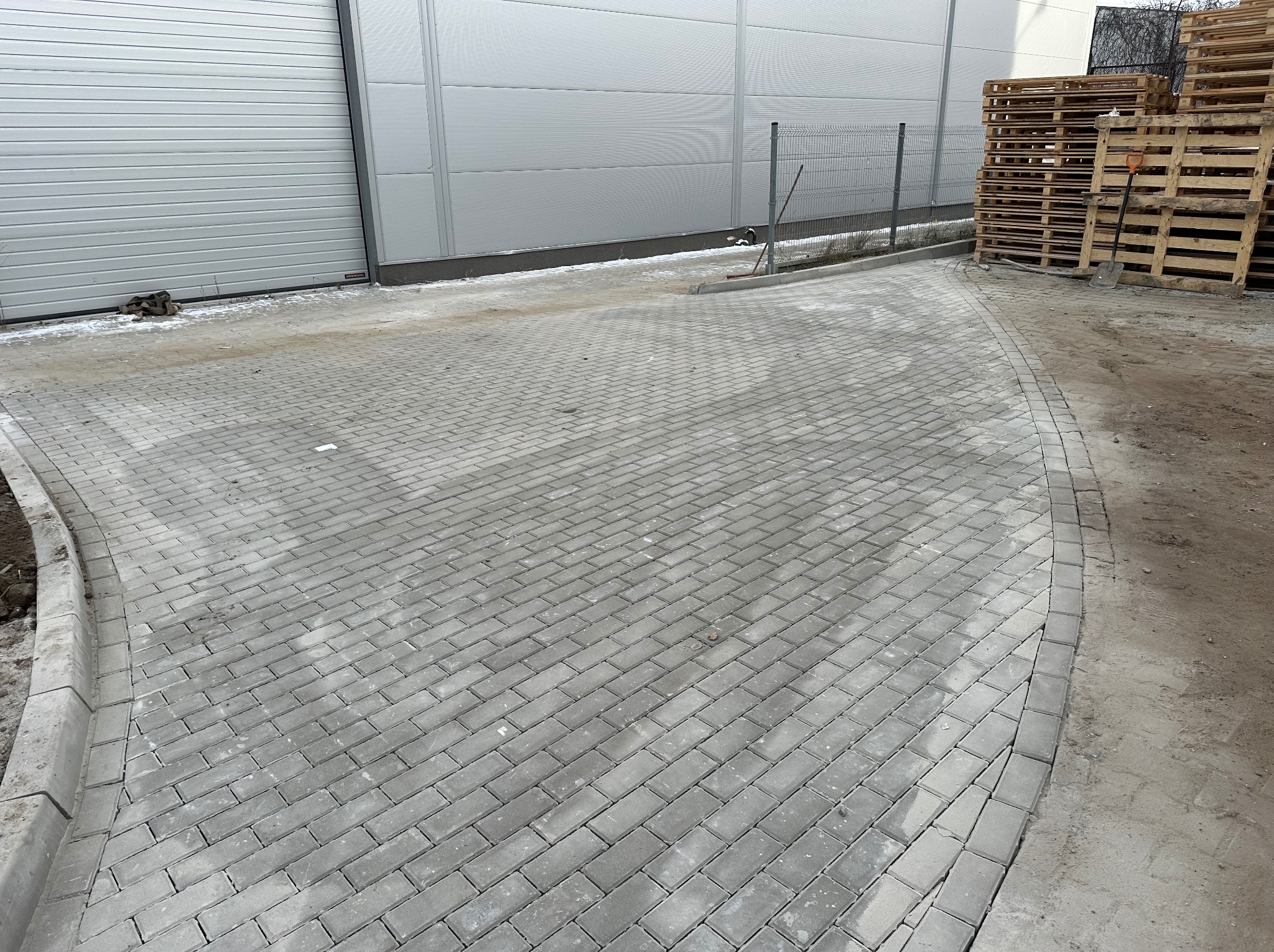 Świeżo ułożona, zakrzywiona ścieżka z szarej kostki brukowej prowadząca obok metalowego magazynu i sterty drewnianych palet.