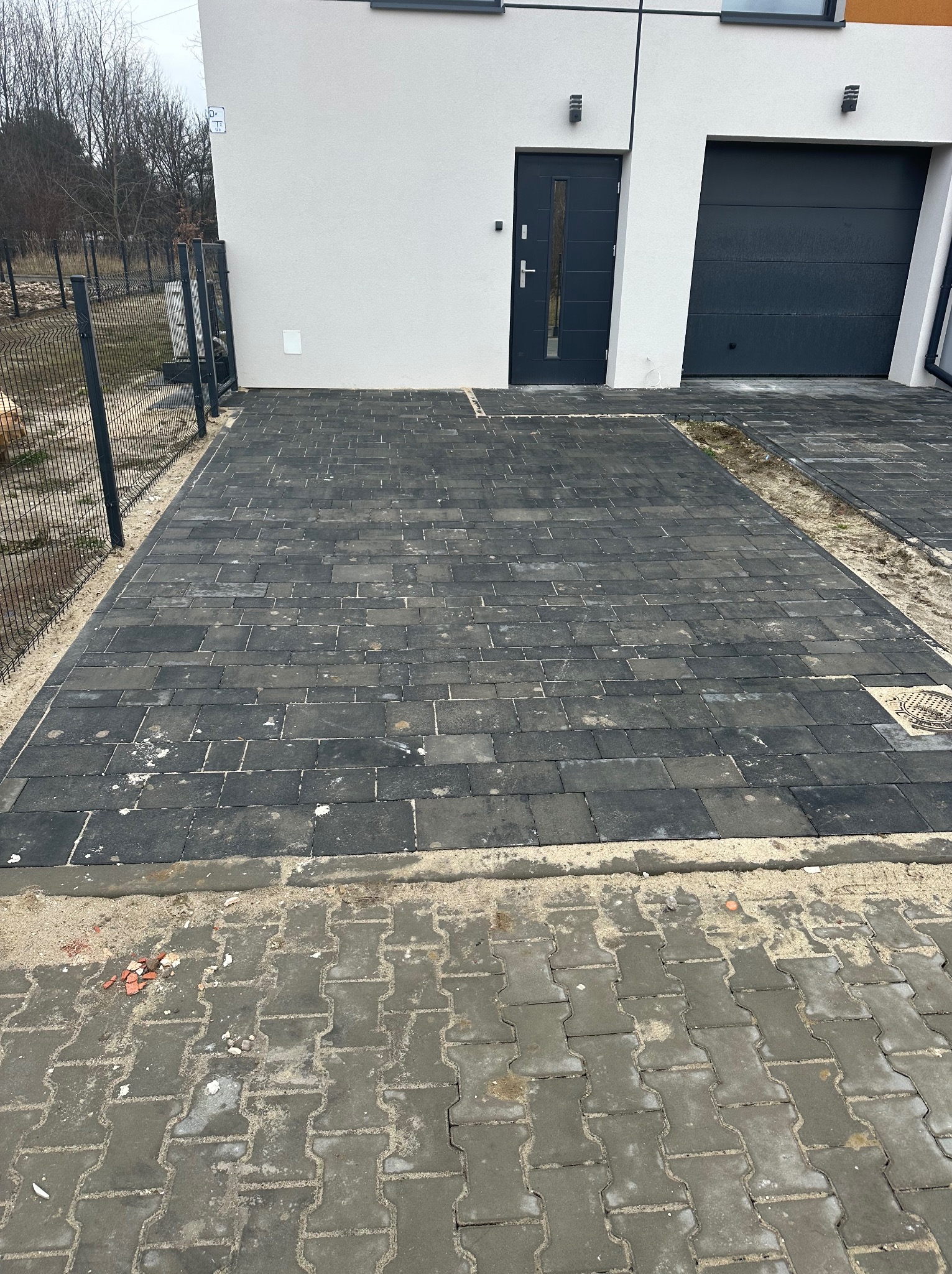 Ułożona, ciemnoszara kostka brukowa przed nowoczesnym domem z szarą stolarką okienną i drzwiową; widoczne fragmenty starej nawierzchni z kostki betonowej oraz pozostałości piasku.