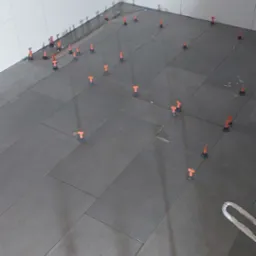 Układanie ciemnoszarych płytek podłogowych w rogu pomieszczenia, z białymi płytkami na ścianach i pomarańczowymi klipsami poziomującymi w spoinach.