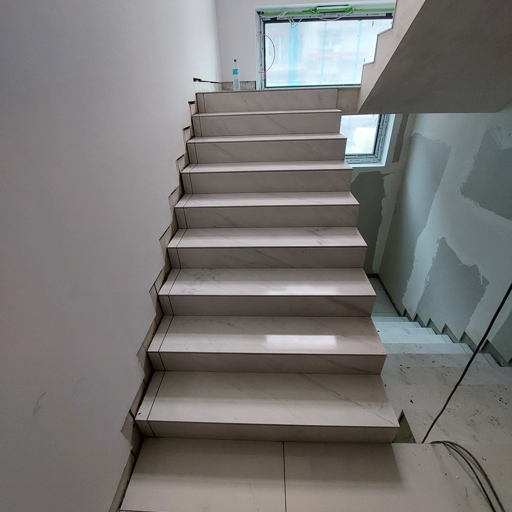 Wykończone schody z białego marmuru z ciemnymi fugami, widoczne niedokończone ściany i zabezpieczone okno w tle.