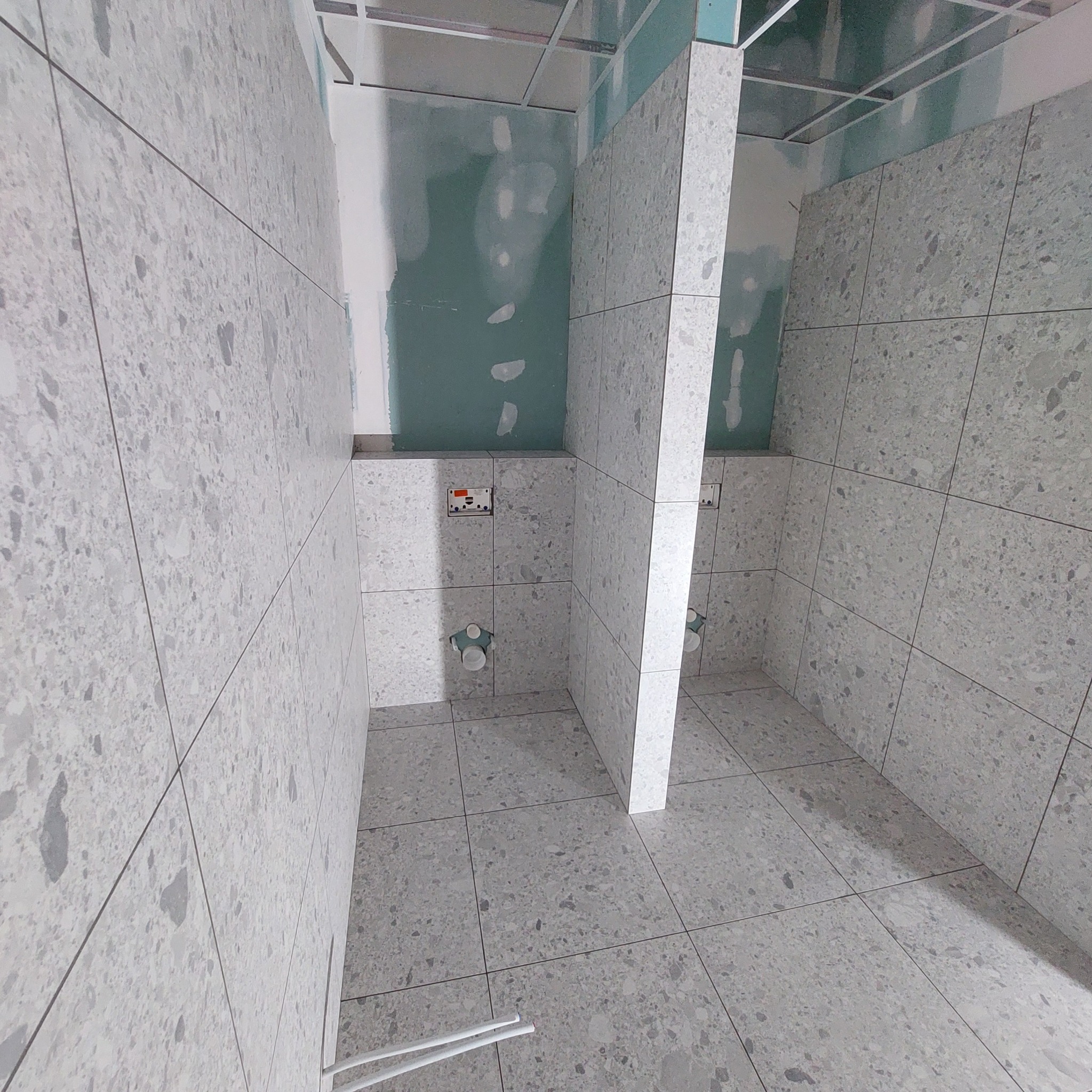 Wnętrze łazienki w trakcie remontu, z widocznymi szarymi płytkami imitującymi kamień na ścianach i podłodze, niezabudowana instalacja wodno-kanalizacyjna oraz płyty kartonowo-gipsowe na suficie.