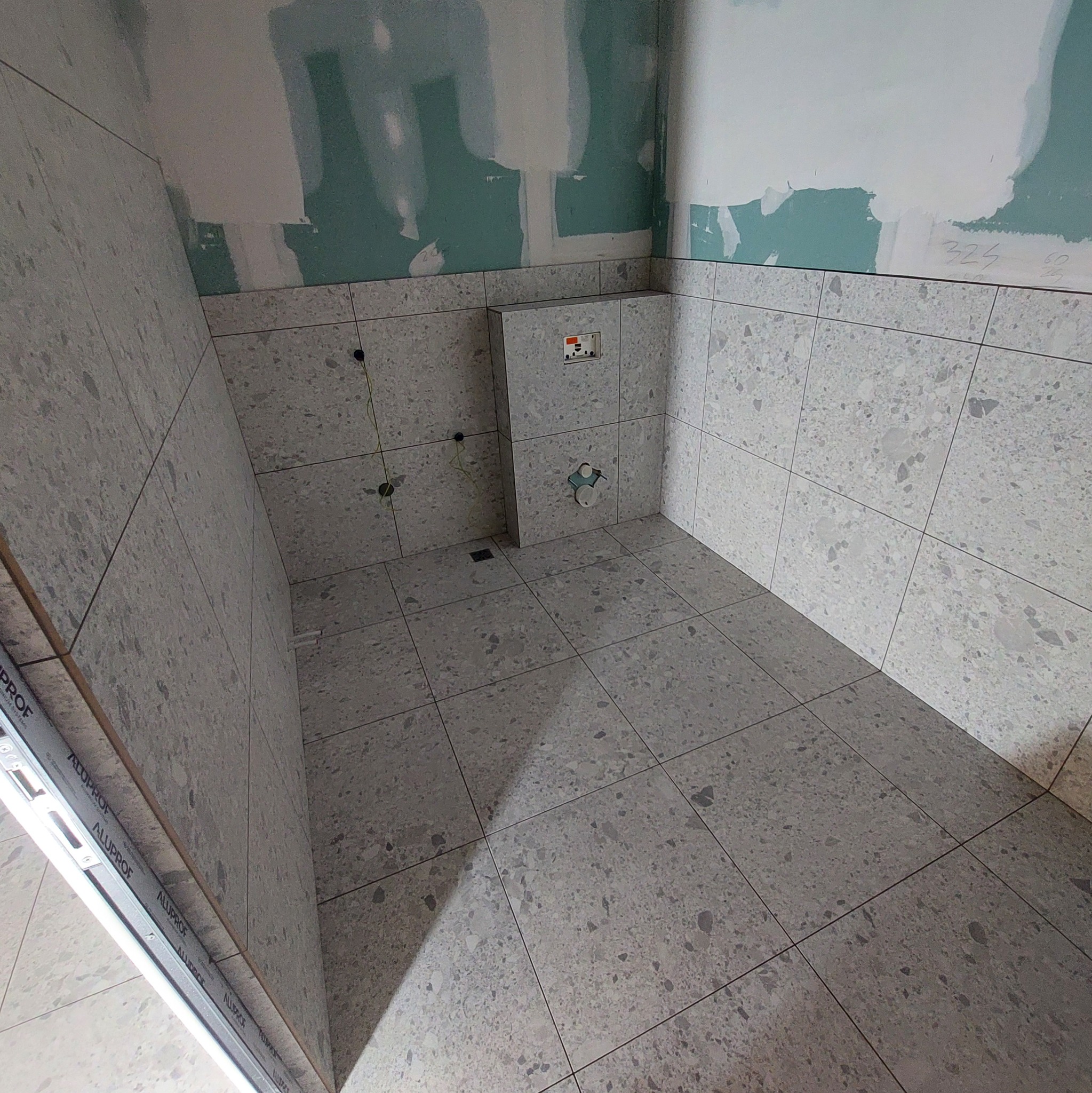 Wnętrze łazienki w trakcie remontu z częściowo ułożonymi szarymi płytkami terazzo na ścianach i podłodze, widoczne otwory na instalacje i fragmenty niezakrytej płyty gipsowo-kartonowej.
