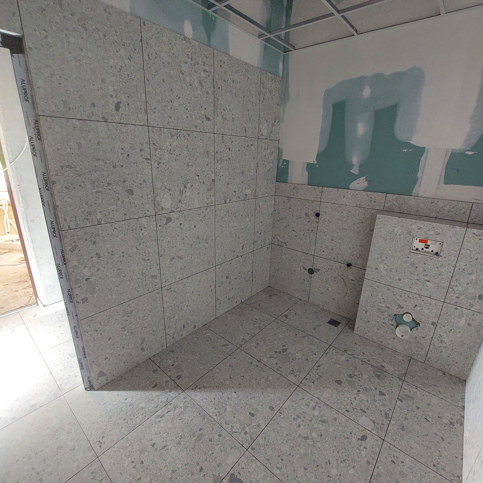 Płytki podłogowe i ścienne z szarego lastryko w łazience w trakcie remontu, widoczne otwory na instalacje i niezakończone prace na ścianach z płyt kartonowo-gipsowych.
