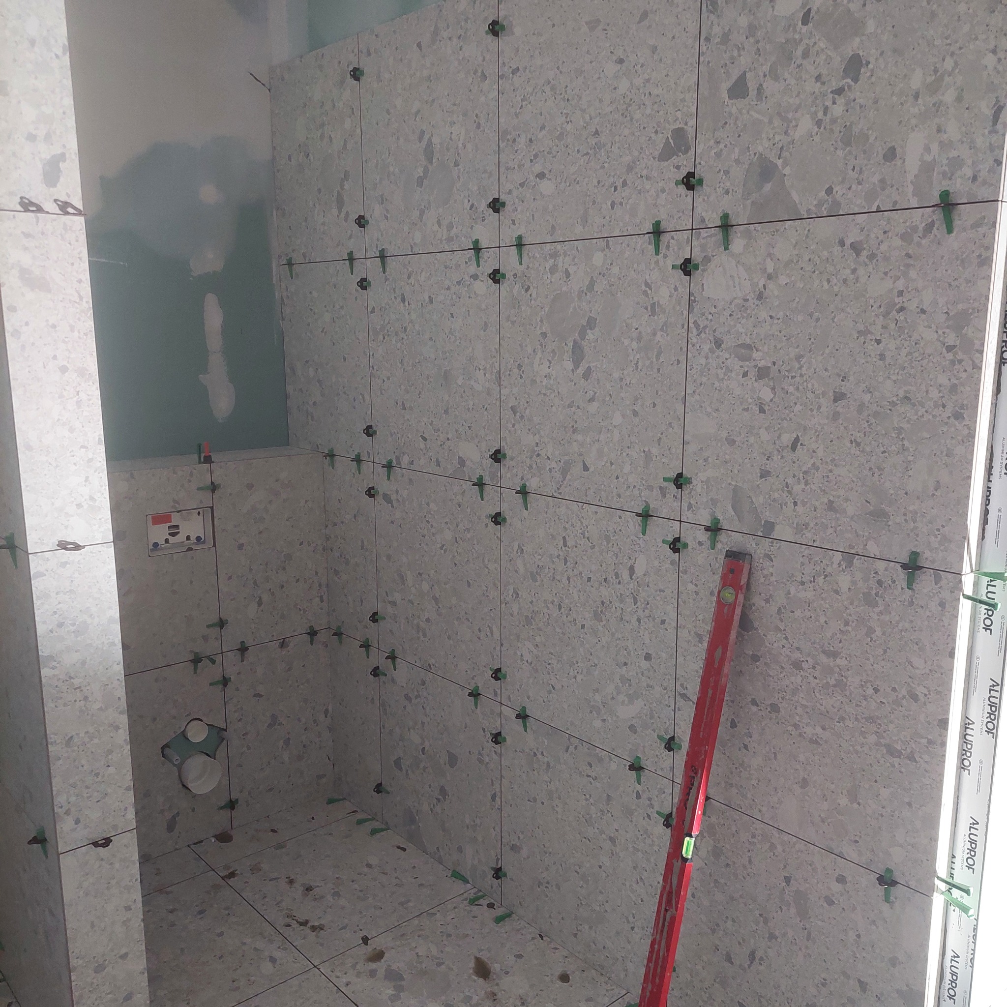 Świeżo ułożone, szare płytki z wzorem lastryko na ścianach i podłodze w trakcie montażu, z widocznymi krzyżykami dystansowymi i czerwoną poziomicą opartą o ścianę.