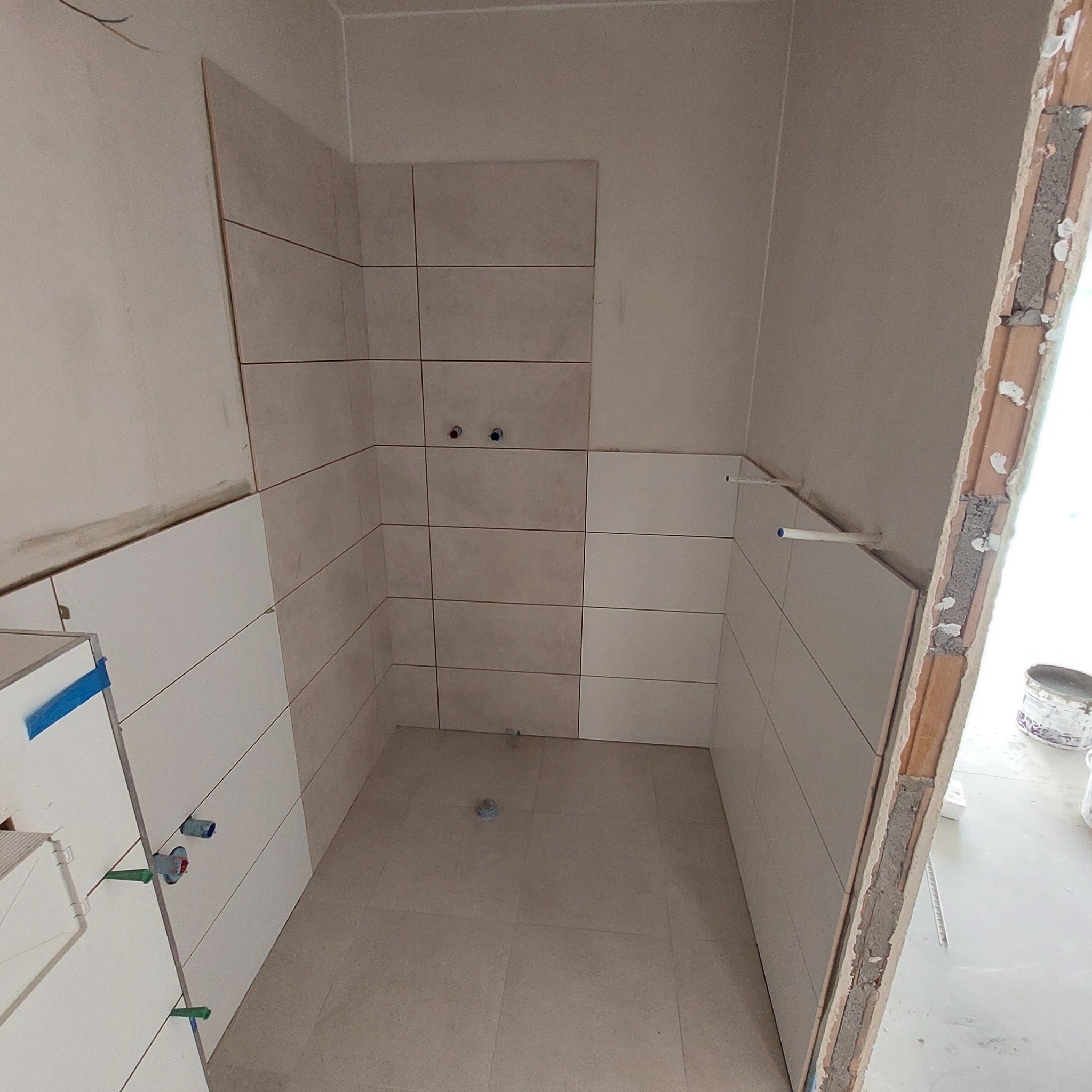 Świeżo ułożone, beżowe płytki w małej łazience w trakcie remontu, widoczne rury i piony wodne, szpary między płytkami, surowe ściany i podłoga, praca w toku.