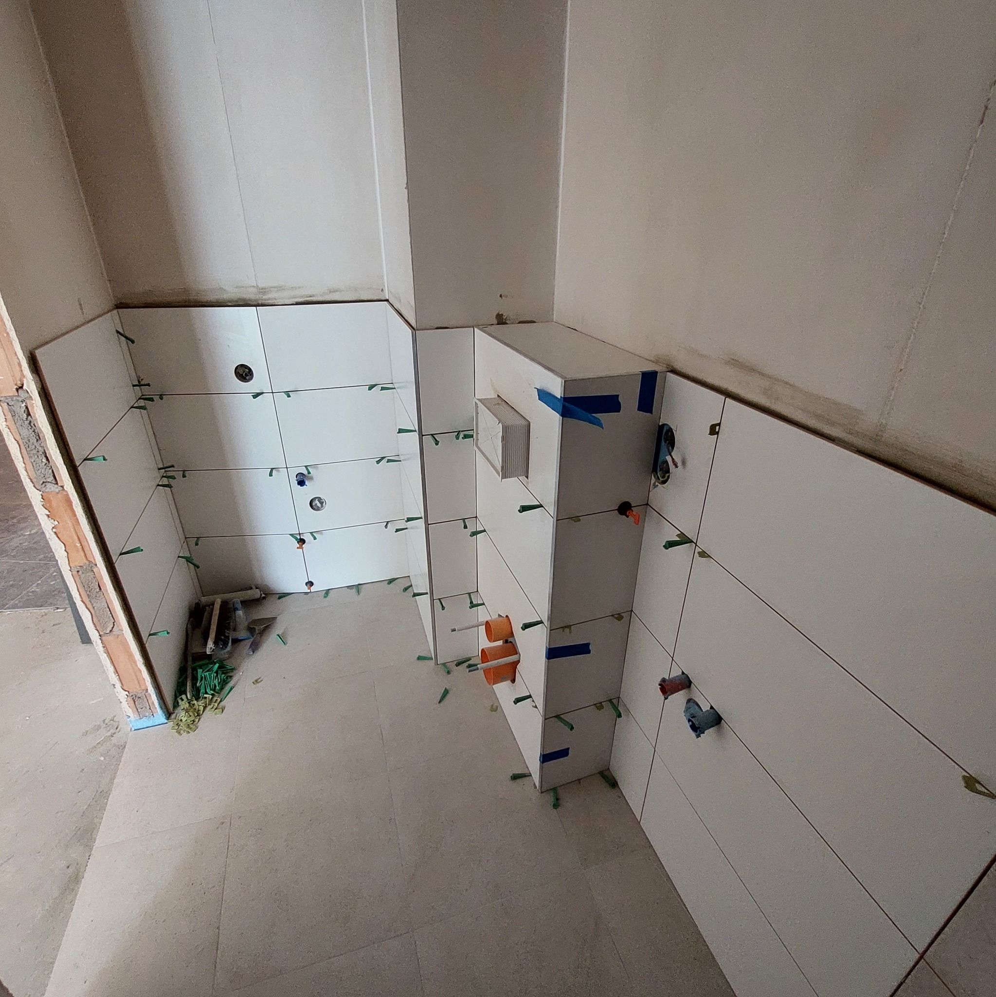 Proces układania białych płytek ceramicznych w pomieszczeniu, widoczne plastikowe krzyżyki dystansowe, fragmenty instalacji wodnej i kanalizacyjnej, oraz niezabezpieczone krawędzie ściany.