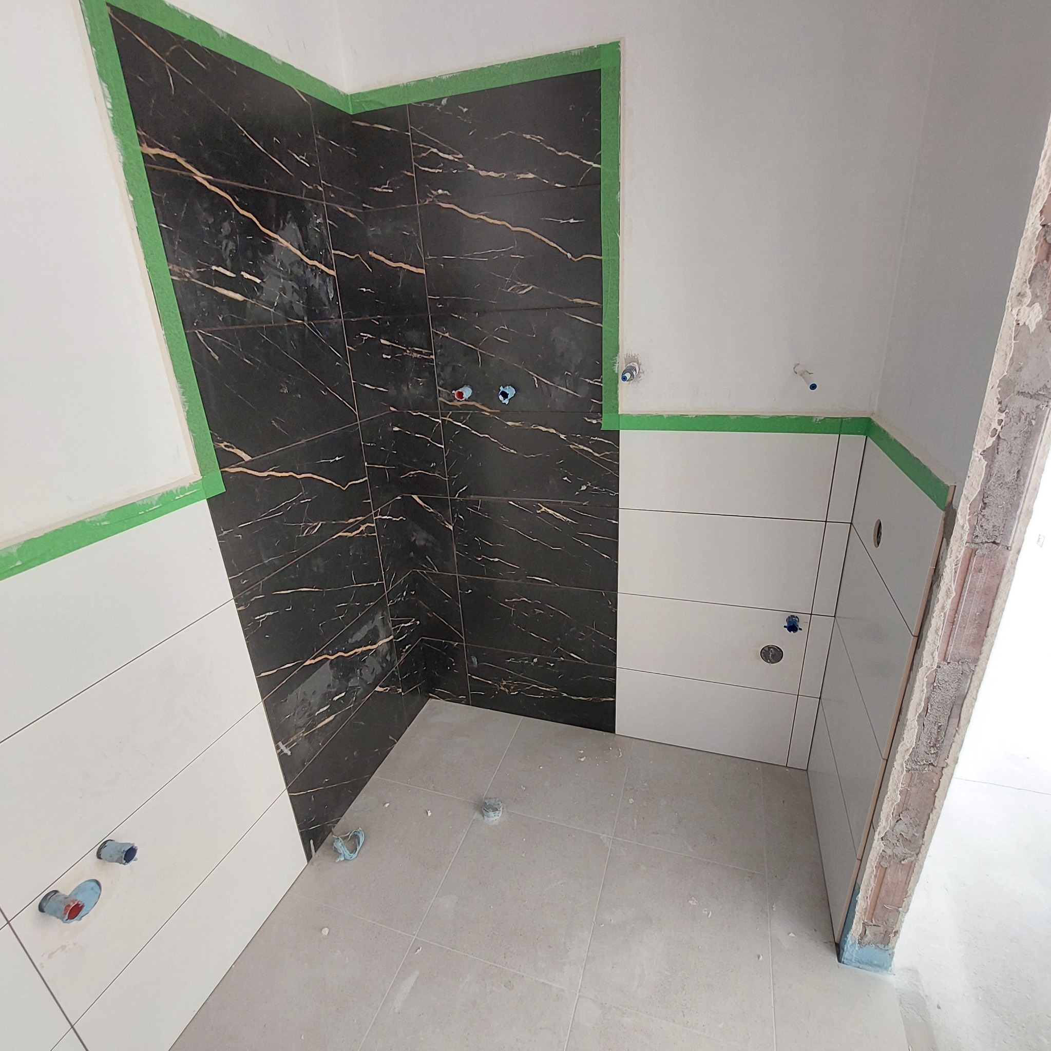 Fragment łazienki w trakcie remontu, widoczne czarne płytki imitujące marmur z wyraźnymi złotymi żyłami, zestawione z białymi płytkami, zabezpieczone zieloną taśmą malarską, z widocznymi...