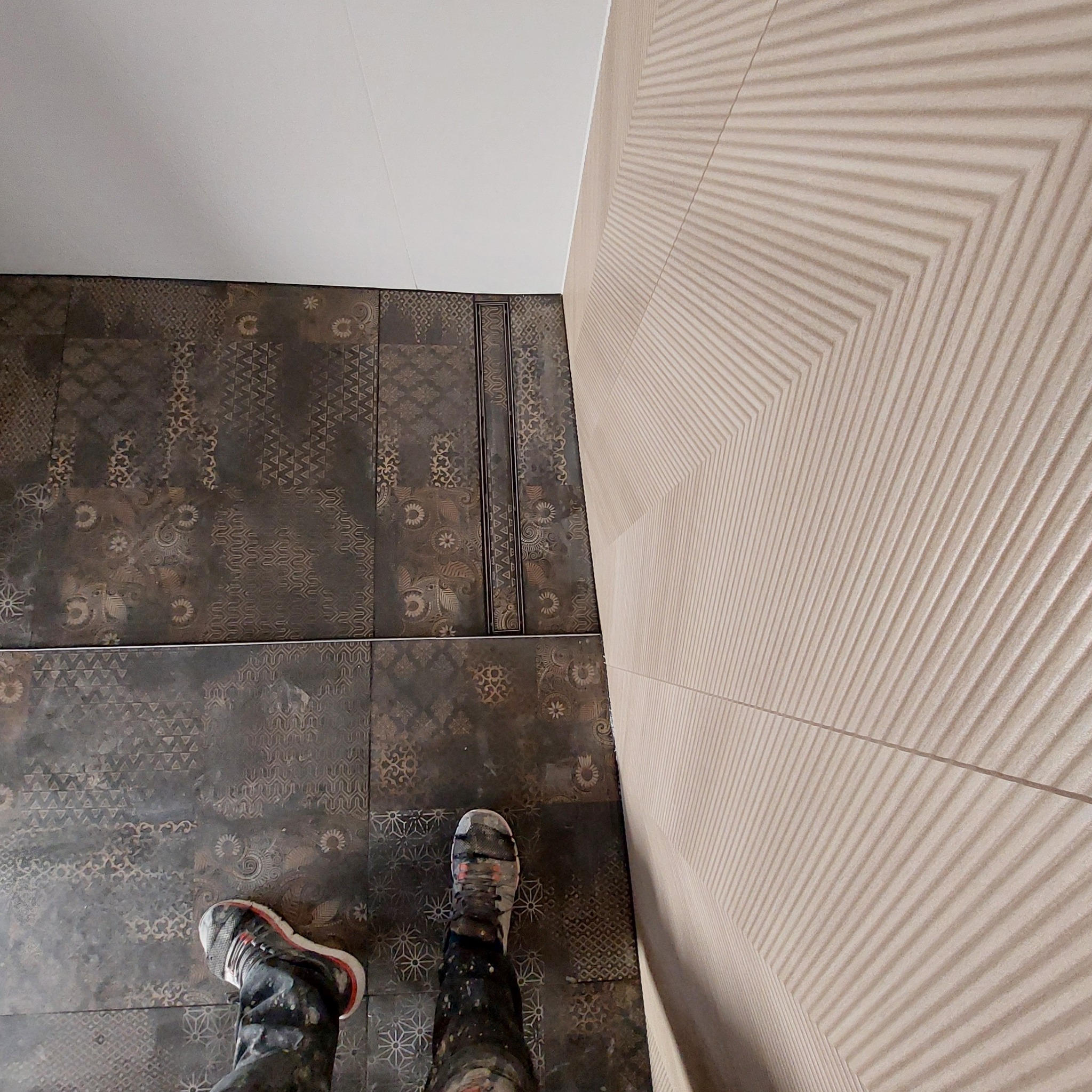 Ułożone płytki podłogowe w ciemnych odcieniach z wzorami i jasne płytki ścienne z liniową strukturą, widok z góry na buty robocze z plamami farby.