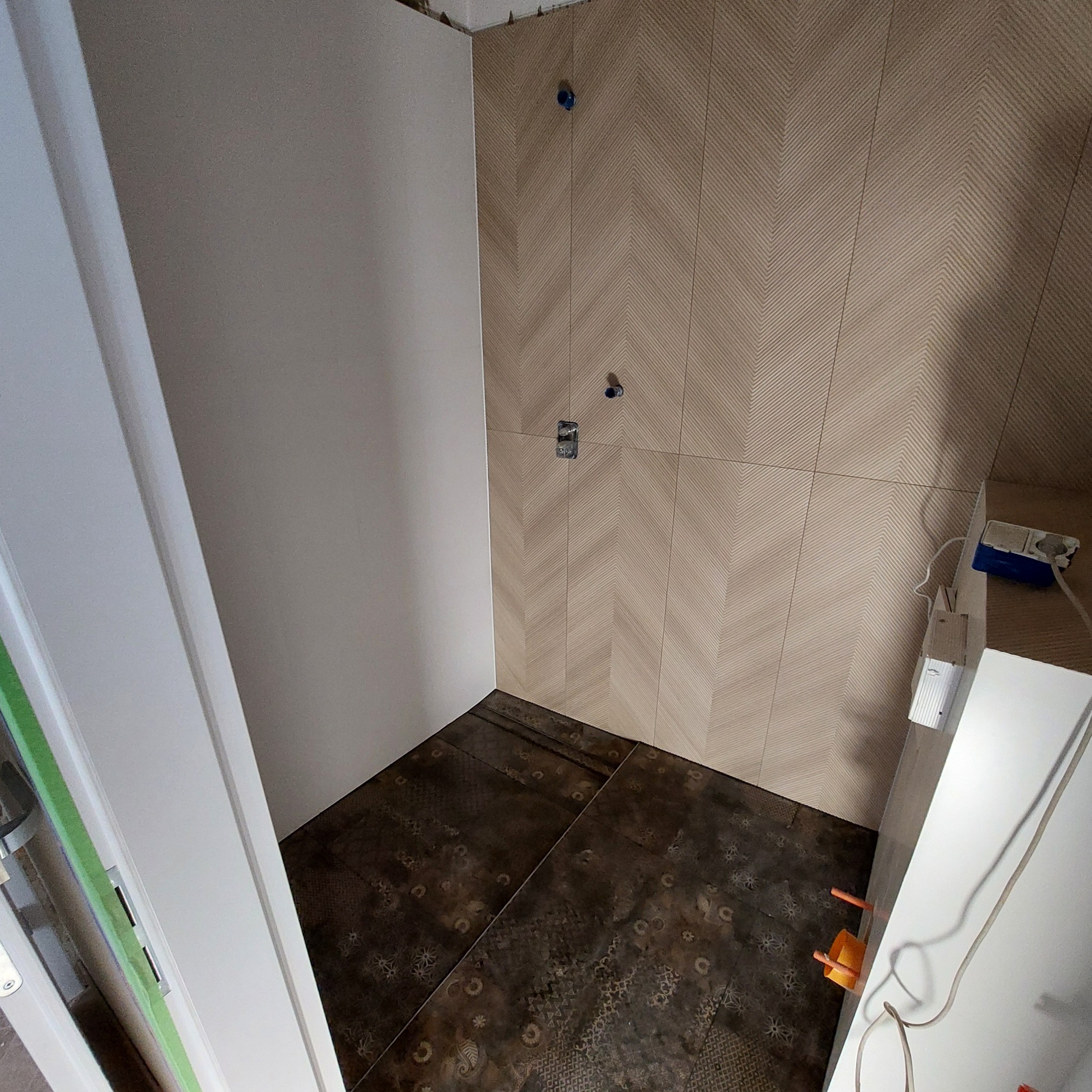 Łazienka w trakcie wykańczania: beżowe płytki ułożone w jodełkę na ścianie, kontrastujące ciemne płytki podłogowe z wzorami, widoczne gniazdka elektryczne i fragment instalacji, surowa ściana...