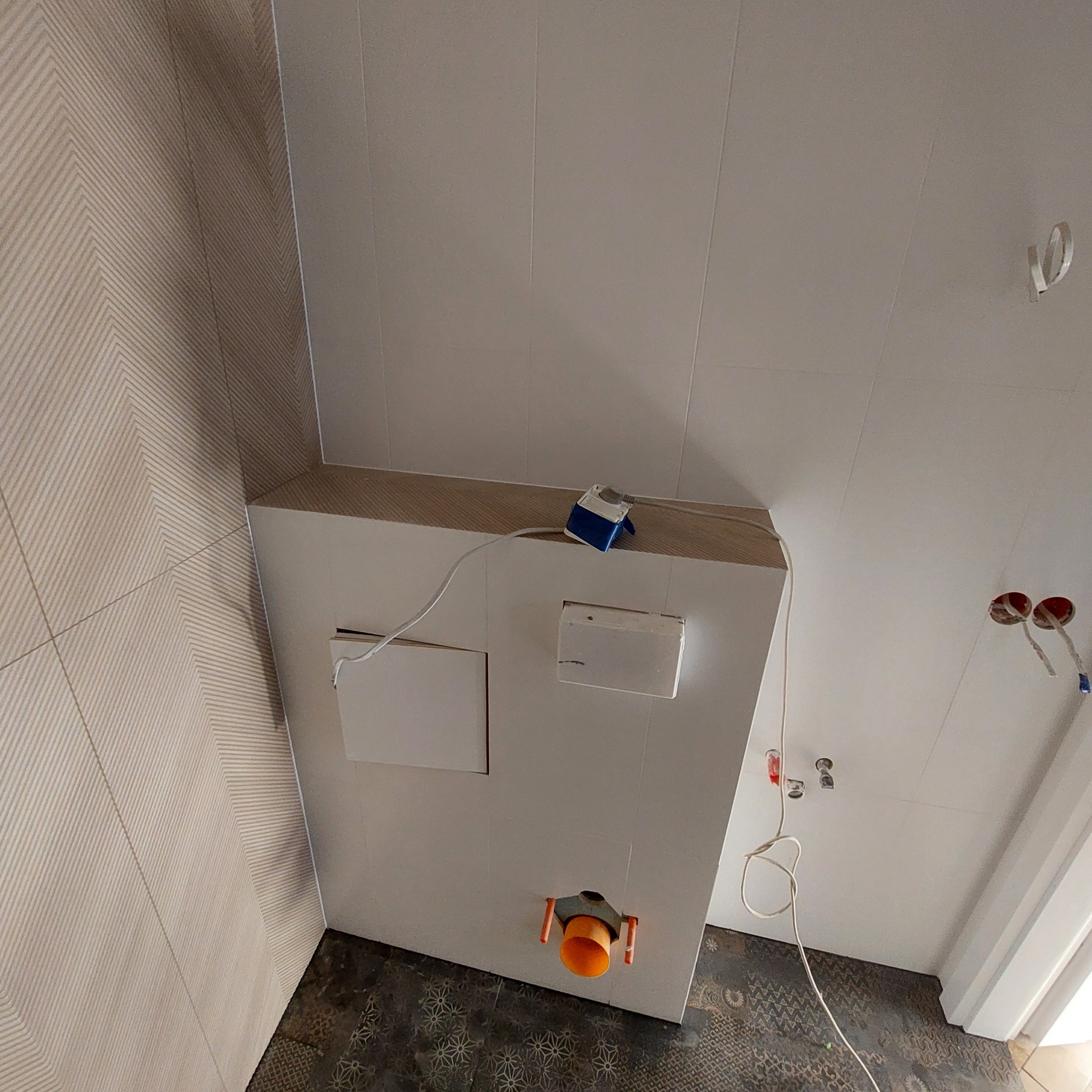 Ściana z zabudową geberit, częściowo wykończona płytkami z delikatnym wzorem, widoczne instalacje wodno-kanalizacyjne i elektryczne, podłoga z ciemnymi płytkami o wzorzystej fakturze.