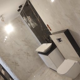 Inwest Dom - Wykończona łazienka z marmurowymi płytkami, wiszącą toaletą i umywalką na szafce. Widoczne czarne akcenty i niedokończone oświetlenie sufitowe.