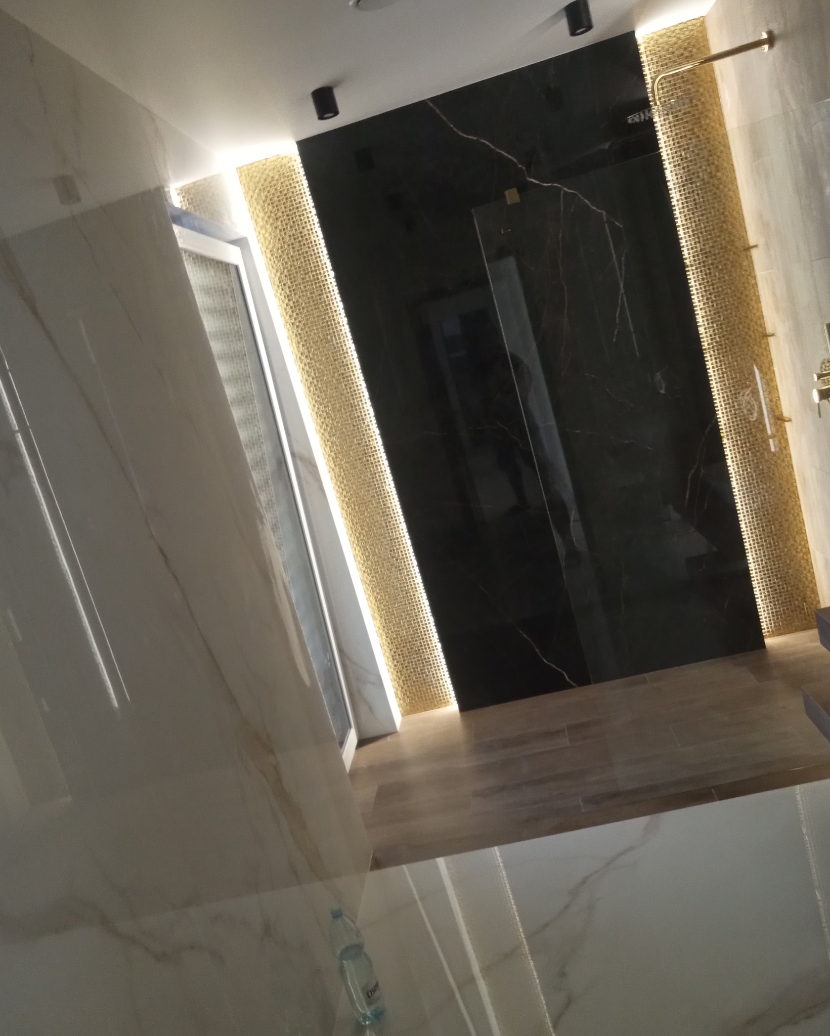 Luksusowa łazienka z czarnymi marmurowymi płytkami, złotą mozaiką i podświetleniem LED, widok z perspektywy podłogi, z butelką wody mineralnej w rogu.