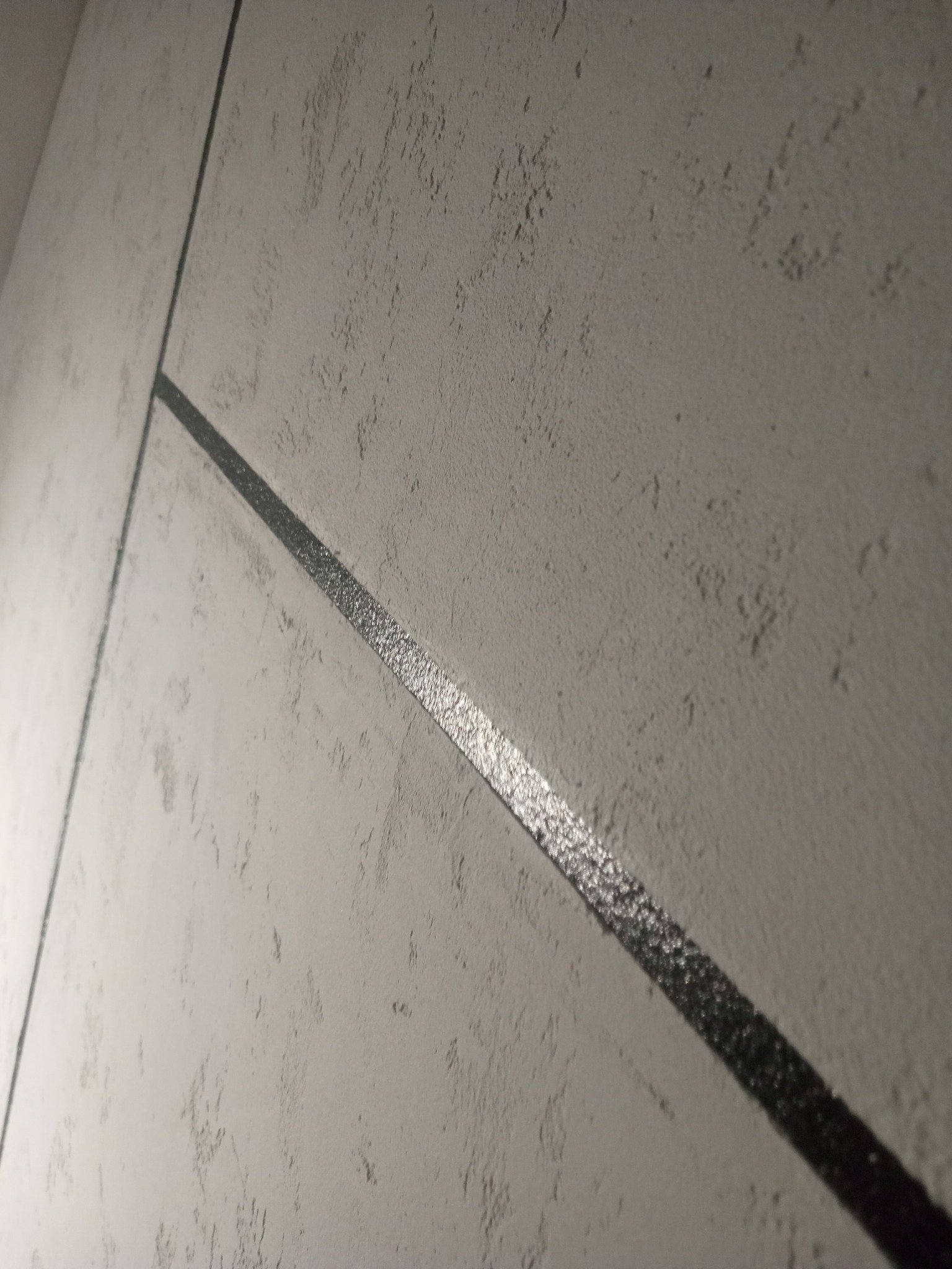 Szara ściana z dekoracyjną, poziomą, ciemną listwą, o strukturze przypominającej tynk strukturalny, oświetlona z góry.