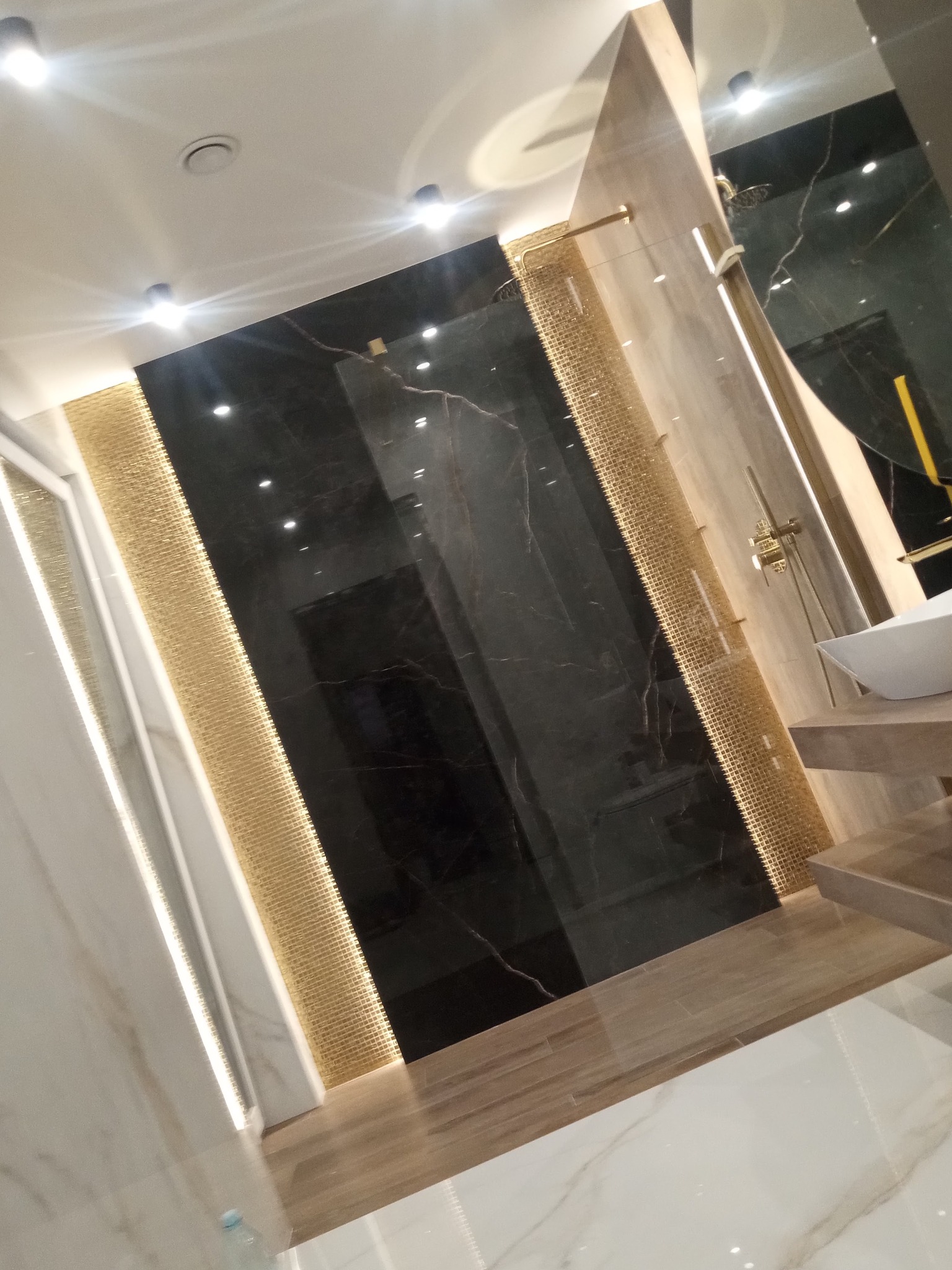 Nowoczesna łazienka z czarnymi marmurowymi płytami, złotymi mozaikami i armaturą w odcieniach złota, oświetlona punktowymi lampami sufitowymi.