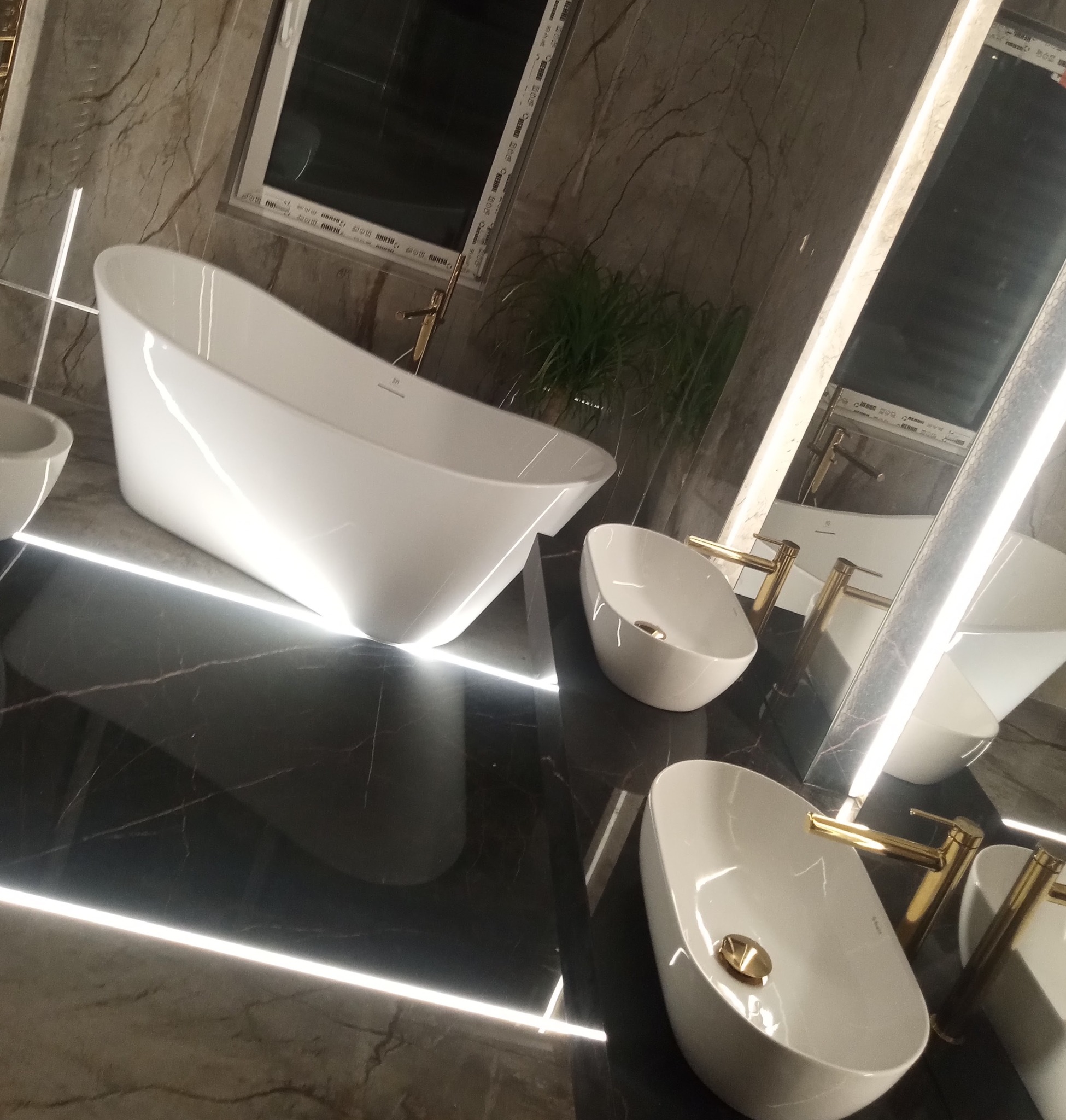Nowoczesna łazienka z wolnostojącą białą wanną, umywalkami nablatowymi z złotymi bateriami na czarnym marmurze z podświetleniem LED, odbicie w lustrze