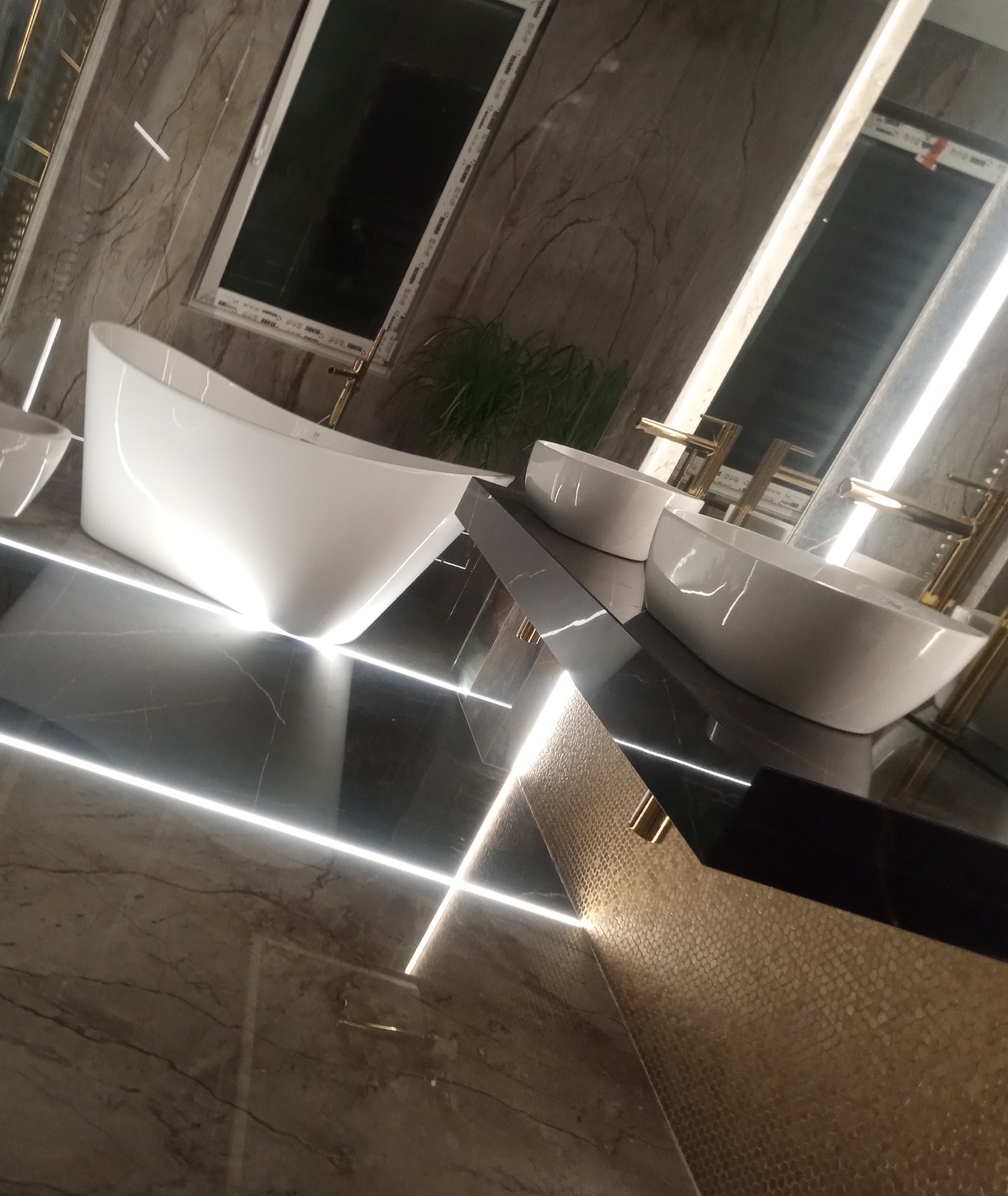 Nowoczesna łazienka z wolnostojącą wanną i umywalkami nablatowymi na czarnym blacie z podświetleniem LED, marmurowe ściany i złota mozaika.