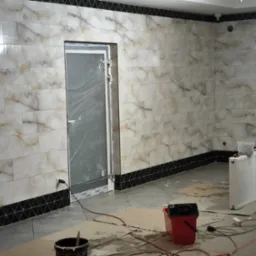 Ściana wyłożona płytkami imitującymi marmur, z czarnym pasem dekoracyjnym u dołu i pod sufitem, drzwi zabezpieczone folią, widoczne narzędzia i akcesoria budowlane sugerujące trwające prace...