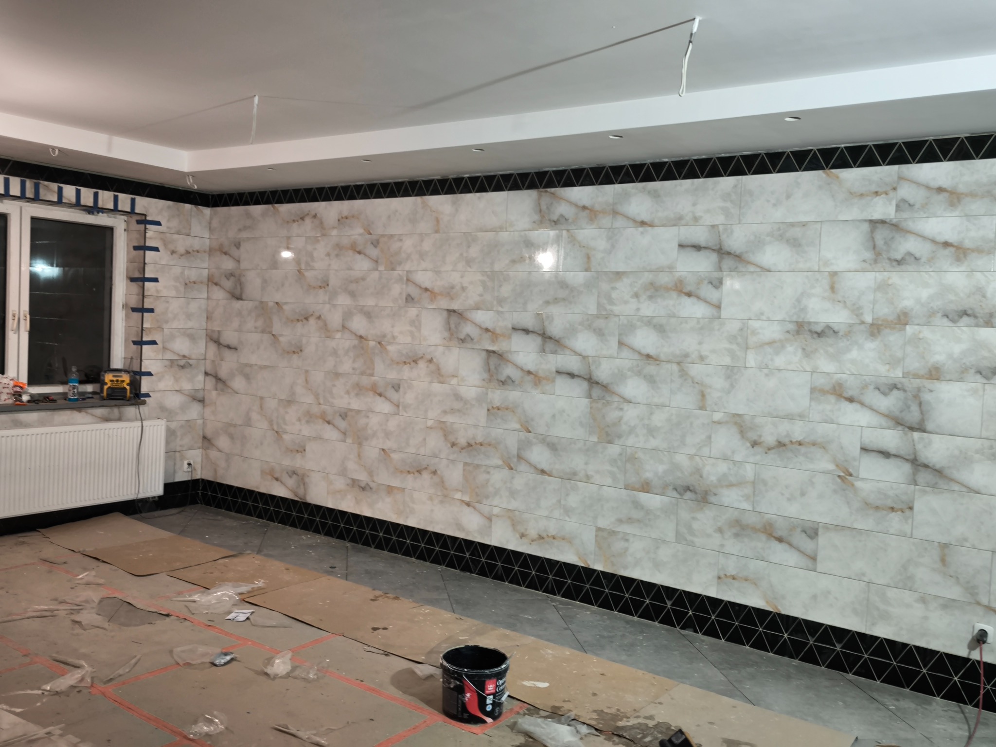 Ściana wyłożona marmurkowymi płytkami z czarnym, trójkątnym wzorem na górze i dole, widoczne narzędzia glazurnicze i wiadro z farbą na podłodze zabezpieczonej kartonem, w trakcie prac wykończeniowych.