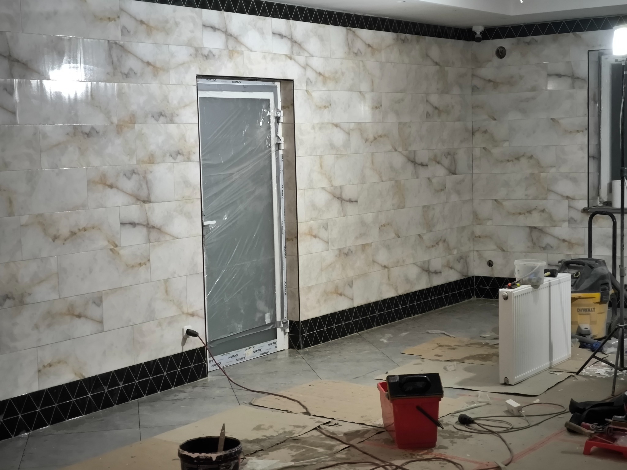 Ściana wyłożona płytkami imitującymi marmur, z czarnym pasem dekoracyjnym u dołu i pod sufitem, drzwi zabezpieczone folią, widoczne narzędzia i akcesoria budowlane sugerujące trwające prace...