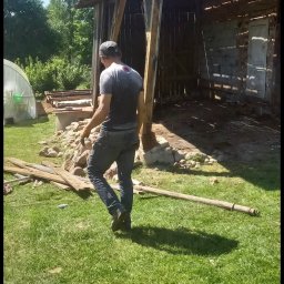 MAJO Piotr Kwiek - Demontaż starej stodoły z ceglanymi ścianami. Mężczyzna w oddali, drewniane elementy konstrukcyjne na trawie. Widoczne pozostałości po budynku.