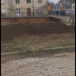 MAJO Piotr Kwiek - Przygotowany teren pod budowę z widocznym budynkiem w tle i przechodzącymi osobami. Ujęcie z poziomu chodnika z kostki brukowej.