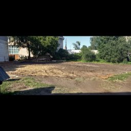 MAJO Piotr Kwiek - Przygotowany teren pod budowę z widocznymi śladami opon, drzewami w tle i częścią budynku. Słoneczny dzień, widok z poziomu gruntu.