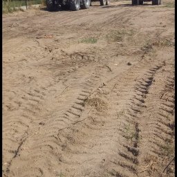 MAJO Piotr Kwiek - Czerwony traktor z przyczepą i koparka na placu budowy. Ślady opon na ziemi, w tle zabudowania i drzewa w słoneczny dzień. Prace ziemne w toku.