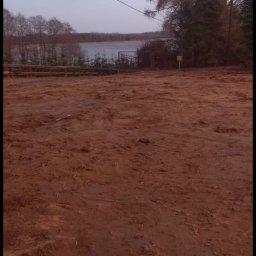 MAJO Piotr Kwiek - Ujęcie terenu po robotach ziemnych z widokiem na jezioro i drzewa. Ślady opon na ubitej ziemi. W tle ogrodzenie i tabliczka ostrzegawcza.