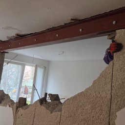 MAJO Piotr Kwiek - Wyburzona ściana z widoczną stalową belką wzmacniającą, podpartą stemplem budowlanym. Fragment ściany z gruzem, okno w tle. Prace wyburzeniowe w trakcie.