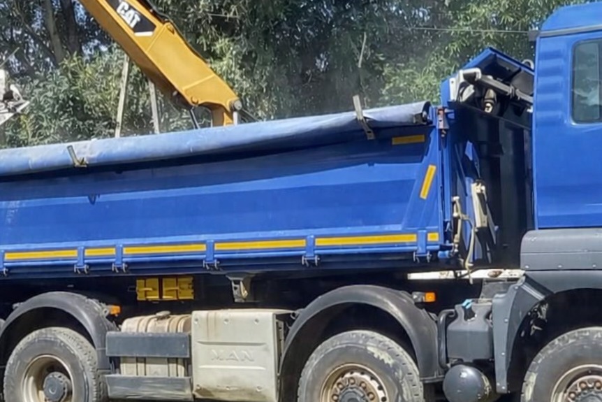 Niebieska wywrotka MAN z żółtym ramieniem koparki CAT podczas załadunku. Widoczne opony, kabina i fragment drzew w tle. Praca w toku, dzień.