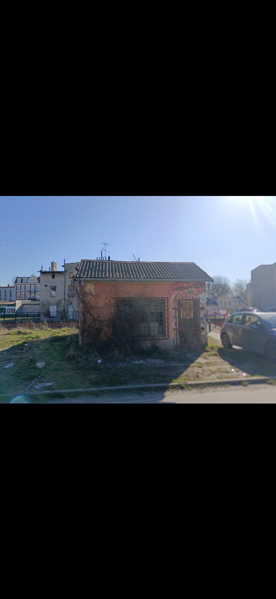 Mały, zaniedbany budynek z graffiti i roślinnością, na tle zabudowy miejskiej w słoneczny dzień. Widoczne ślady rozbiórki i zaniedbania.