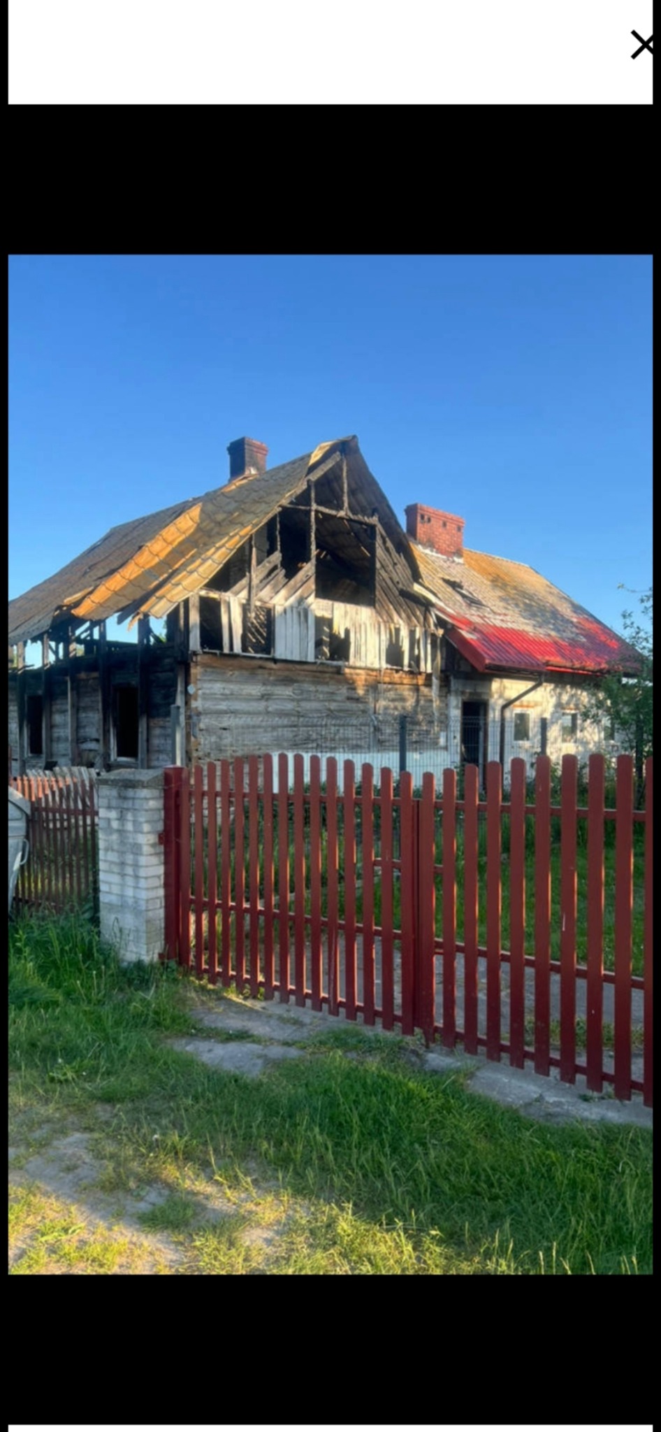 Zniszczony, drewniany dom z częściowo zawalonym dachem i wypalonymi fragmentami, otoczony czerwonym płotem. Widoczne ślady pożaru i dewastacji.