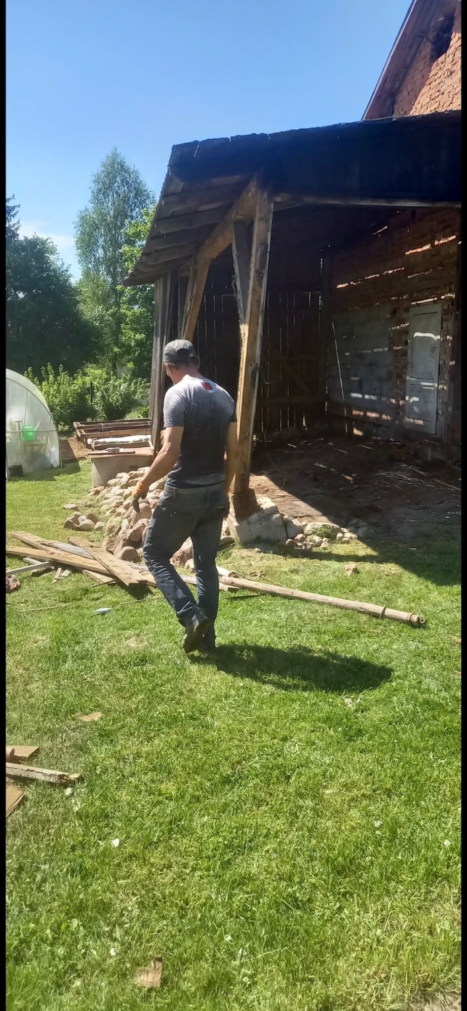 Demontaż starej stodoły z ceglanymi ścianami. Mężczyzna w oddali, drewniane elementy konstrukcyjne na trawie. Widoczne pozostałości po budynku.