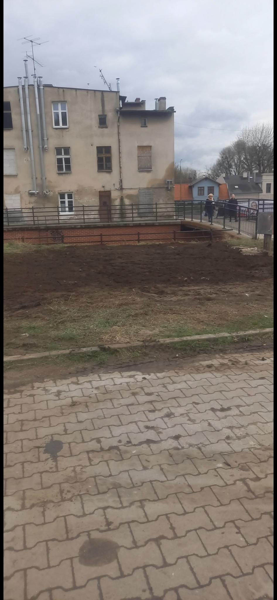 Przygotowany teren pod budowę z widocznym budynkiem w tle i przechodzącymi osobami. Ujęcie z poziomu chodnika z kostki brukowej.