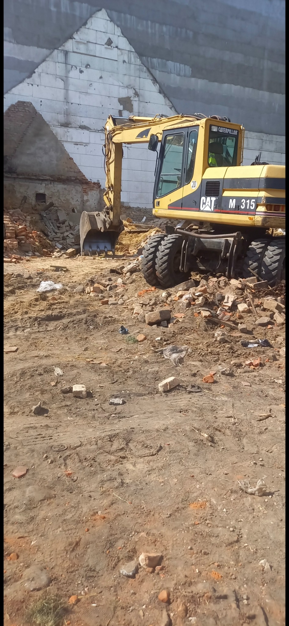 Żółta koparka CAT M 315 na placu rozbiórkowym w Ornecie, gruz i cegły w tle. Prace rozbiórkowe w toku, widoczne ślady opon na ziemi.