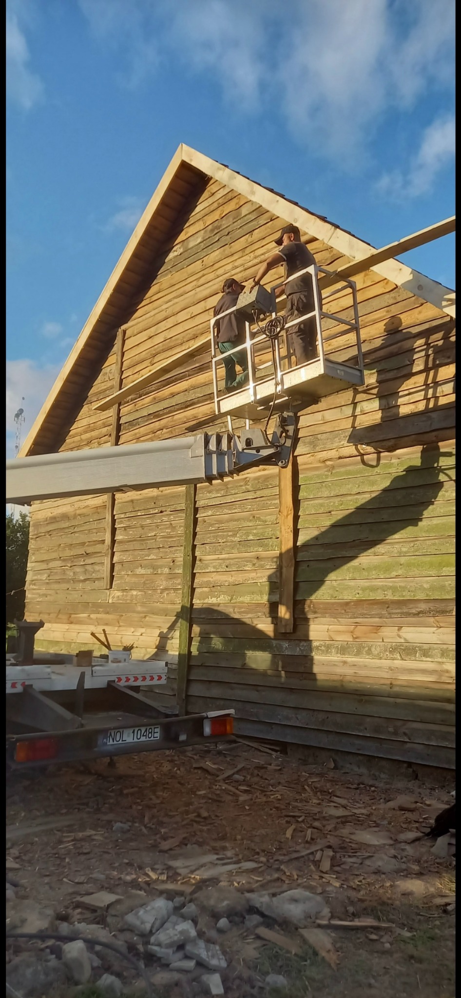 Montaż drewnianej konstrukcji budynku z podnośnika koszowego. Dwóch pracowników w trakcie pracy na tle błękitnego nieba. Widoczny fragment pojazdu z podnośnikiem.